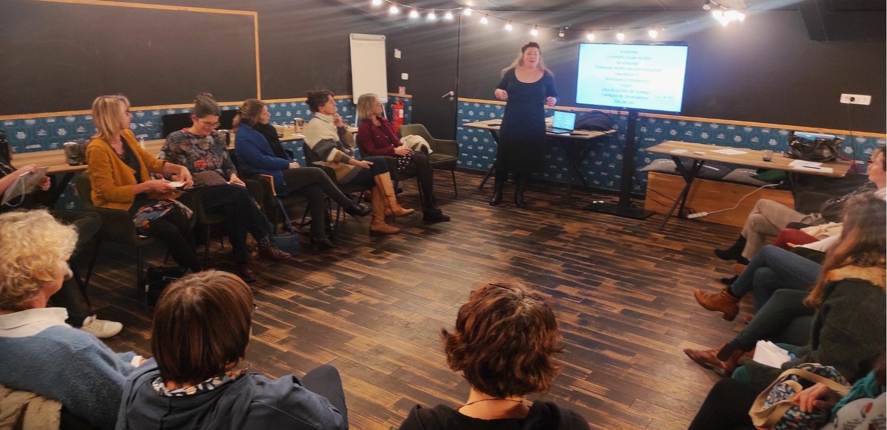 Une femme donne une présentation devant un groupe de personnes assises dans une salle sombre, avec un écran numérique à l'arrière et un tableau blanc à côté.