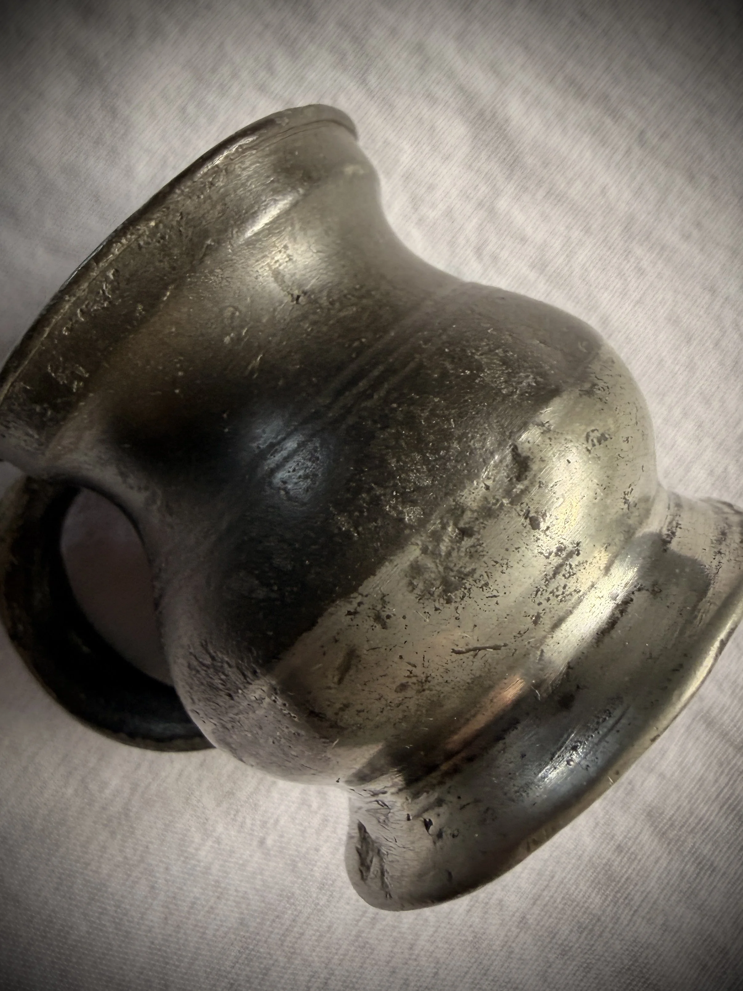 Close-up of a weathered antique pewter cup with a deep grey finish, showing small dents and signs of historical use on a linen surface.