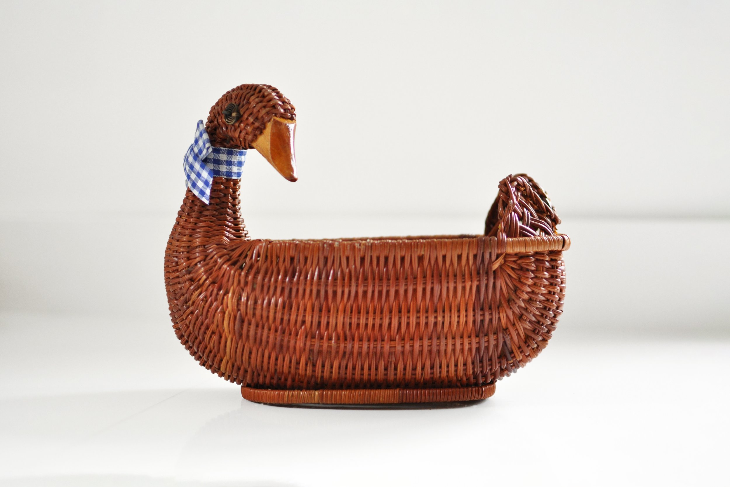 A vintage wicker duck-shaped basket with a blue checkered ribbon around its neck, set against a plain white background.