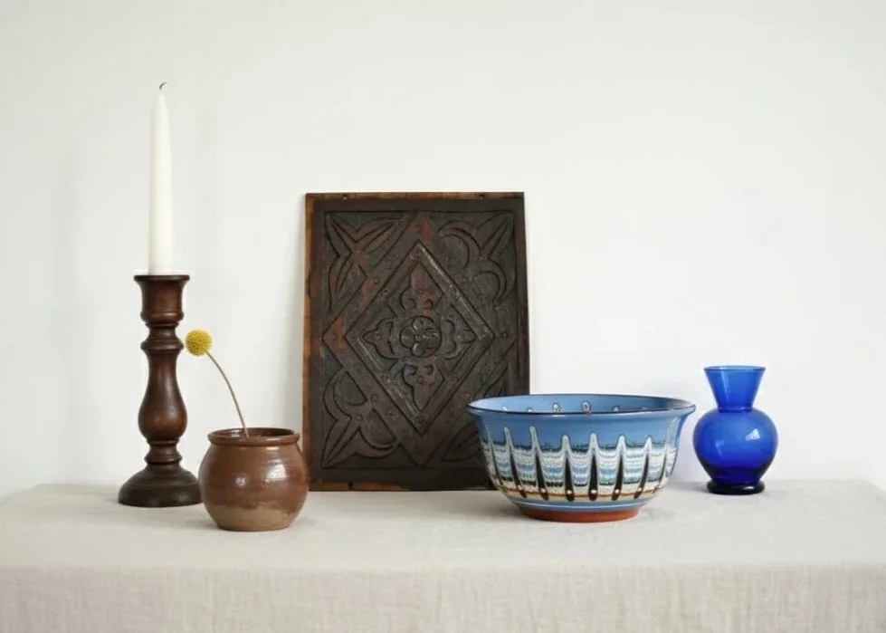 A table with a white cloth displaying a brown candlestick with a white candle, a small brown pot with a yellow flower, a square dark wooden wall art piece, a large colorful ceramic bowl, and a small cobalt blue vase.