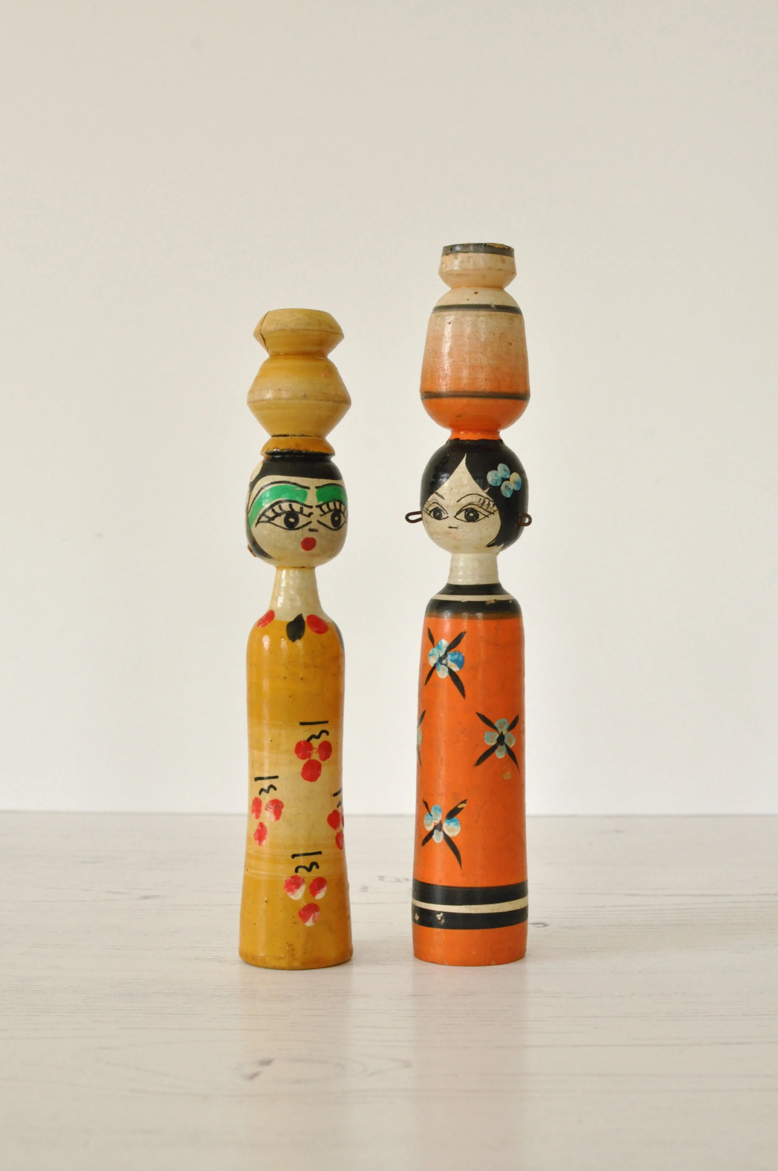 Two painted wooden figurines with black hair and floral designs, each with a stack of smaller painted wooden pieces balanced on their heads, standing on a white surface against a plain background.