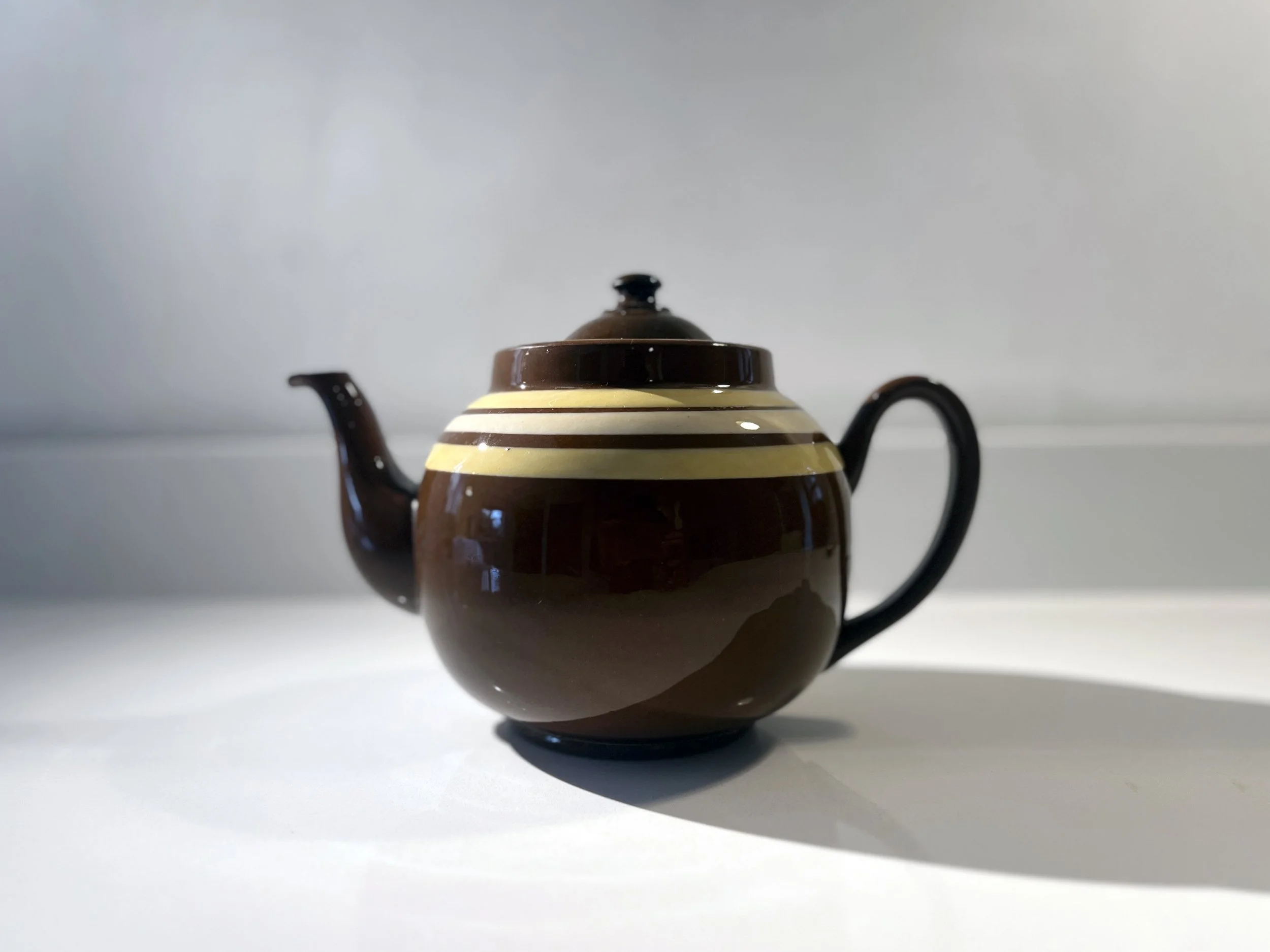 A small, round, brown ceramic teapot with a black handle and lid, decorated with three cream-colored horizontal stripes around its upper part, sitting on a plain white surface.