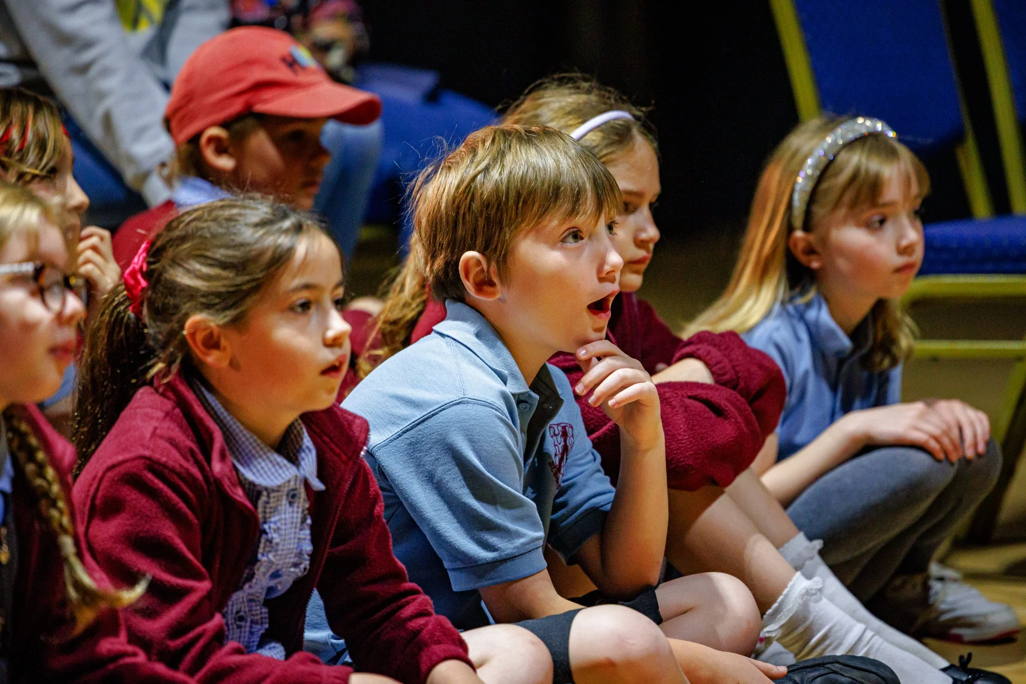 Four children sit cross-legged watching a performance. Their mouths are open as they watch.