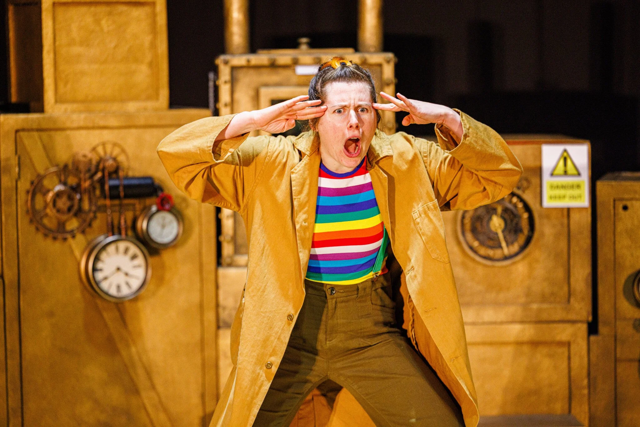 A woman with a brown coat and rainbow striped top holds her hands to her head and stares in amazement