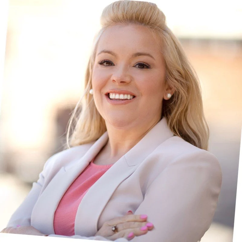 Smiling person with blonde hair wearing a light blazer and pink top, arms crossed.