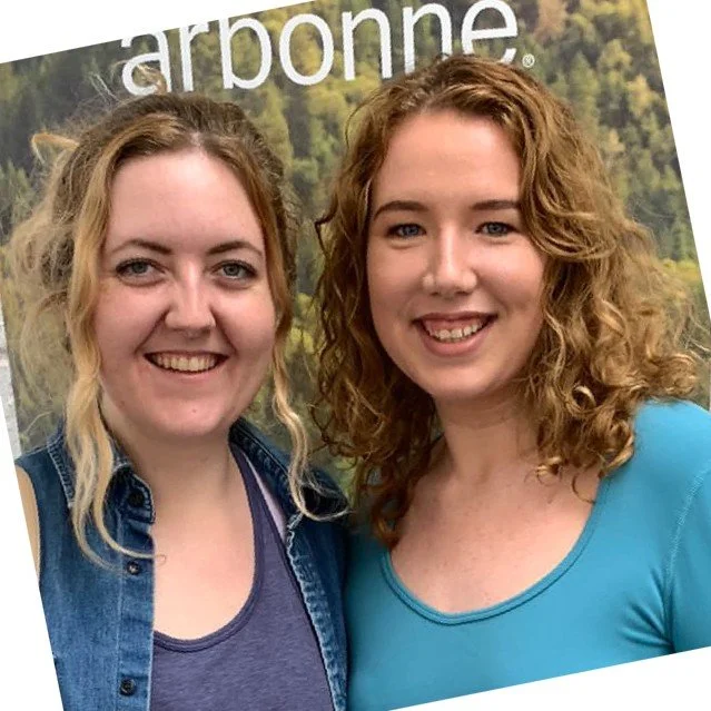 Two women smiling in front of a forest-themed background with the word "arbonne."