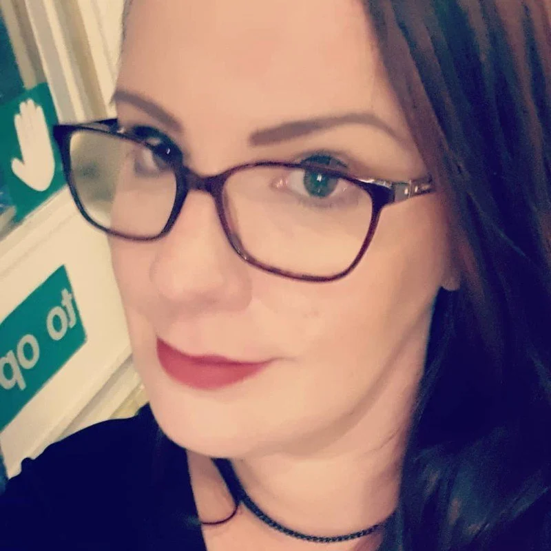 Close-up of a woman with glasses and red lipstick, indoors, with a blurred sign in the background.