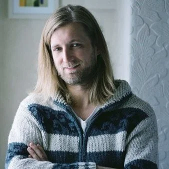 Person with long blond hair wearing a striped sweater, smiling with arms crossed.