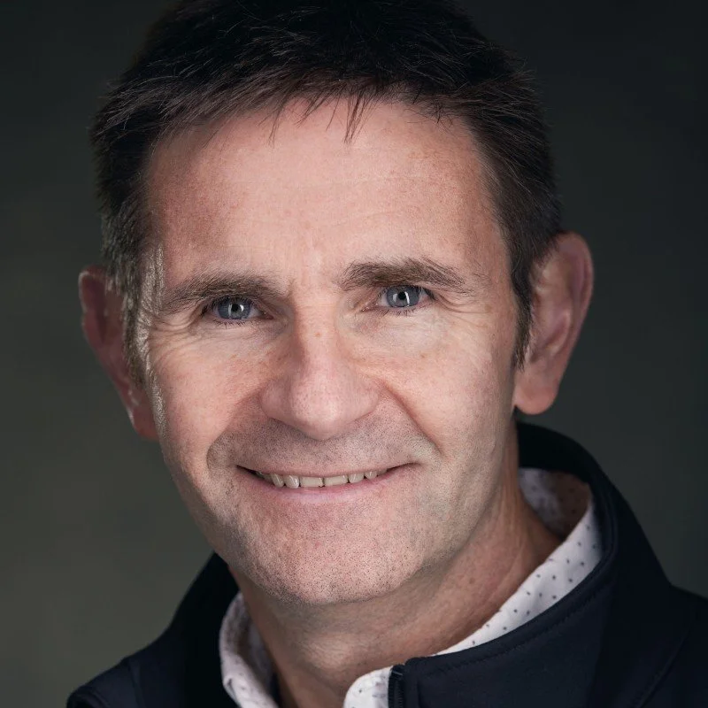 Portrait of a middle-aged man smiling, wearing a dark jacket and light-colored shirt, with a neutral background.