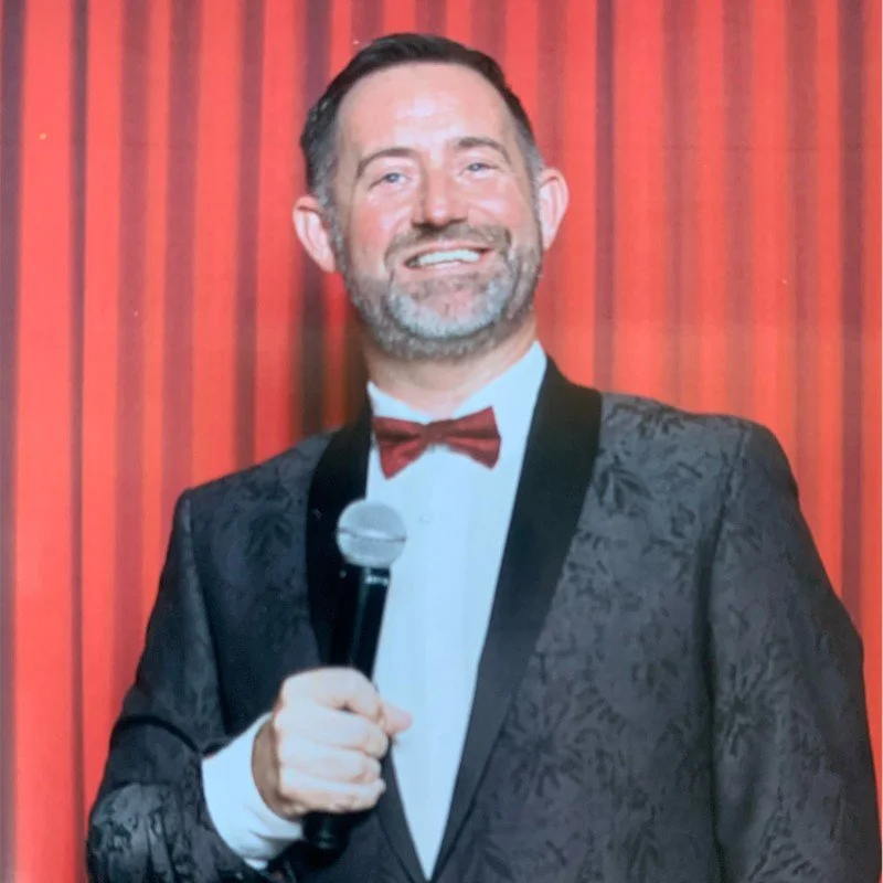 Man in a patterned tuxedo and red bow tie holding a microphone, standing in front of a red curtain background.