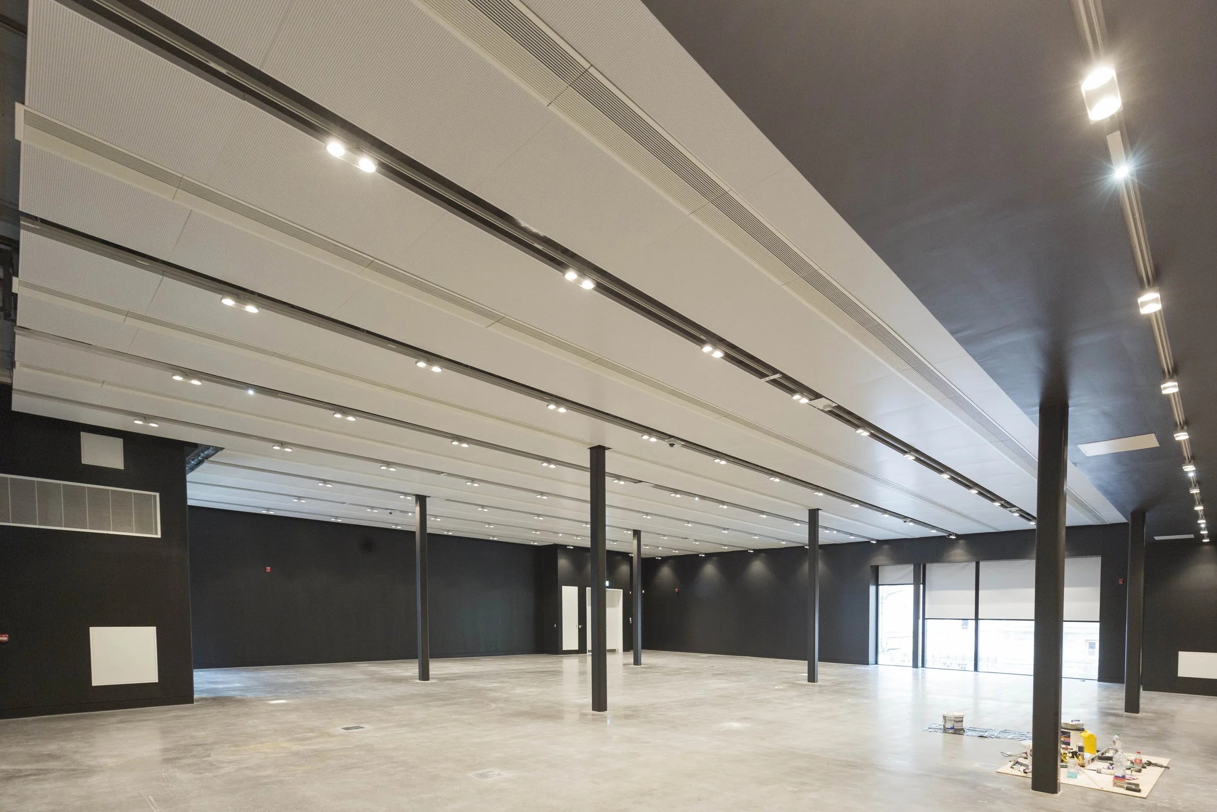 Large empty room with black walls, gray ceiling panels, and multiple ceiling lights, supported by black metal columns, with construction materials on the floor.