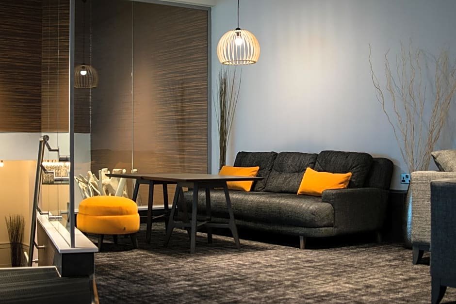 Modern living room with a gray sofa, yellow cushions, a round yellow ottoman, a wooden coffee table, pendant light, and decorative branches in a minimalist setting.