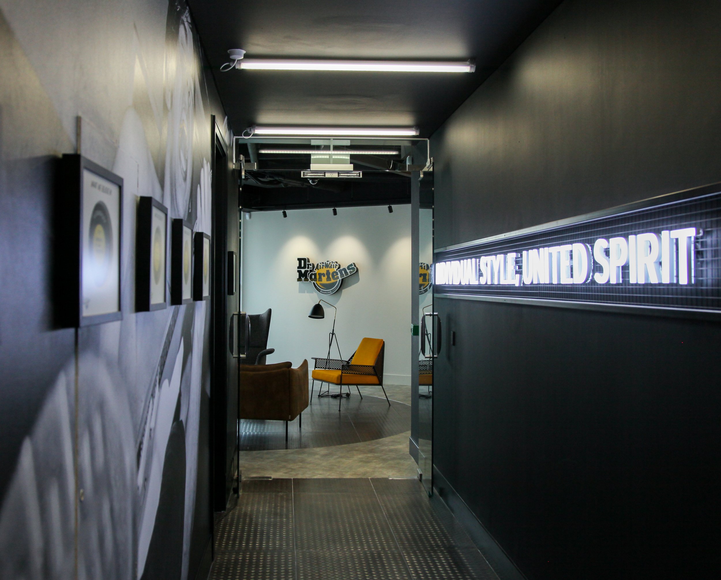 Modern office hallway leading to a lounge area with armchairs and a lamp, featuring wall art and a sign that reads 'Individual Style, United Spirit', with a logo in the background.
