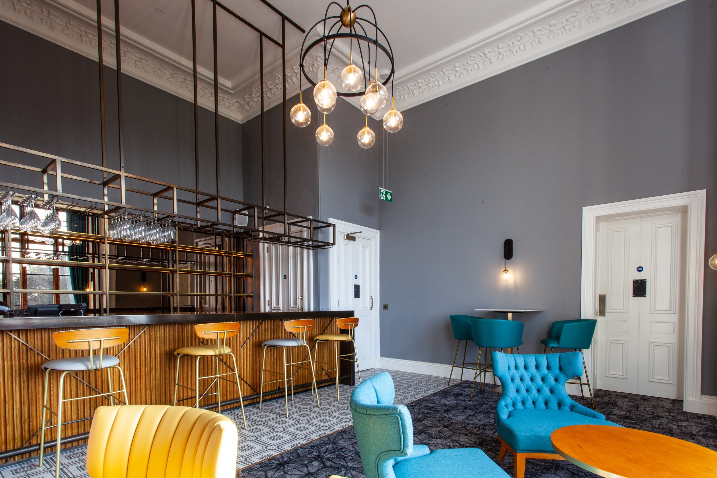 Modern bar area with high ceilings featuring a long wooden bar counter and yellow bar stools, overhead glass storage, blue and yellow lounge chairs, and a round table with teal chairs. A chandelier with glass bulbs hangs from the ceiling.