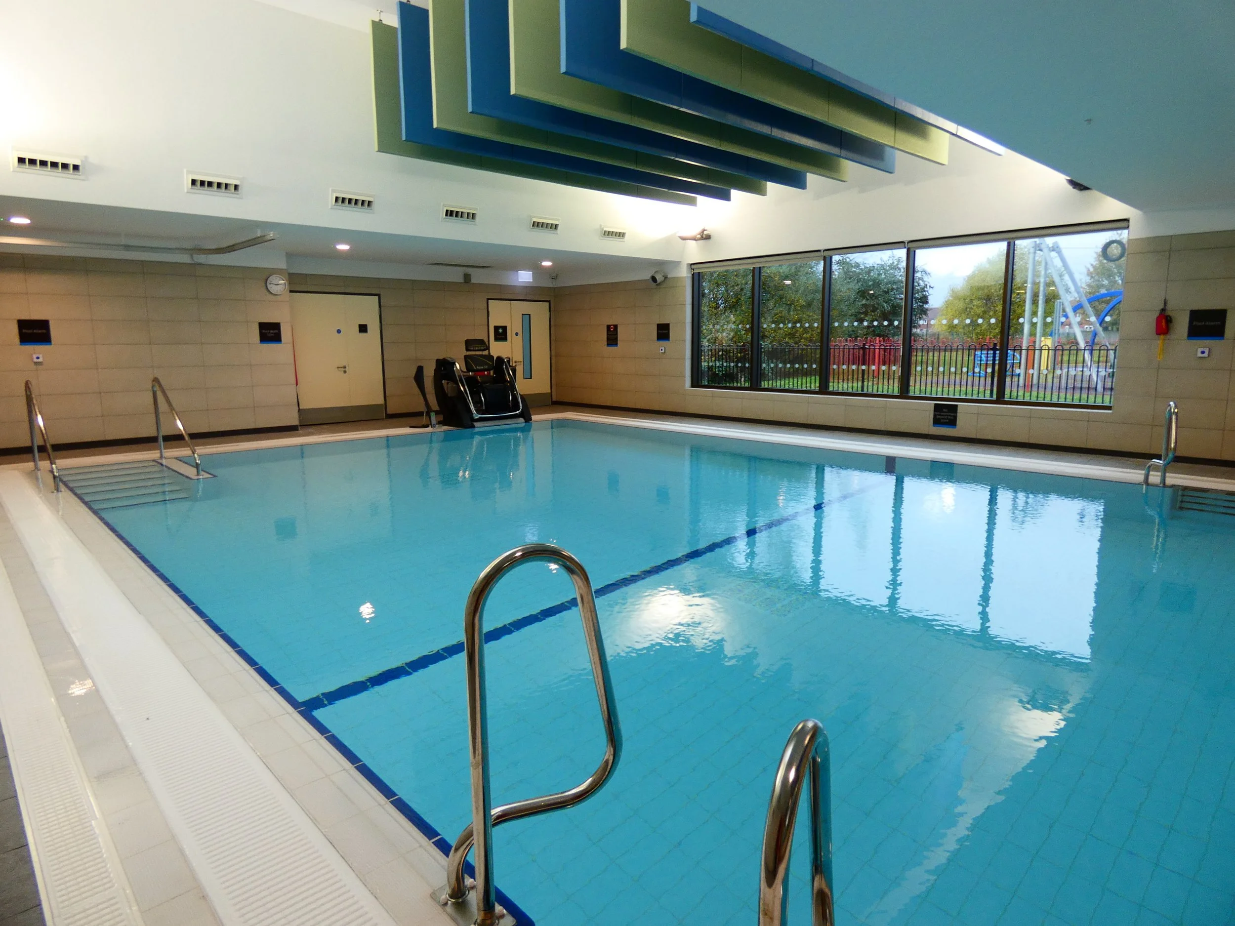 Indoor swimming pool with blue water and stainless steel handrails, surrounded by beige walls. Large windows on one side show an outdoor view with trees and a playground. Ceiling features geometric designs in shades of blue and green.