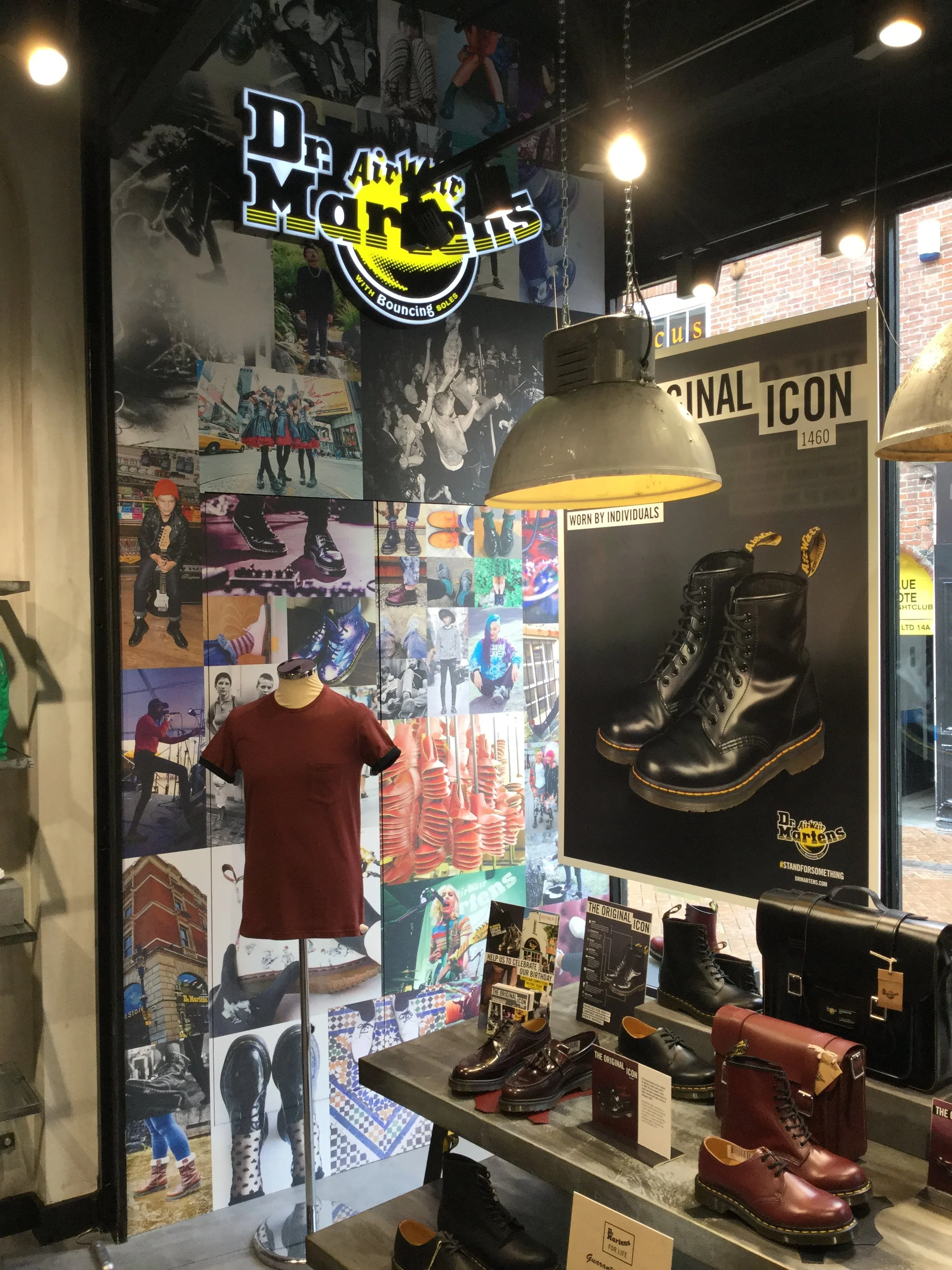 Dr. Martens retail store display featuring various footwear and accessories, including signature boots and shoes. The backdrop showcases a collage of images and a brand logo, with promotional signage highlighting the "Original Icon 1460." A mannequin