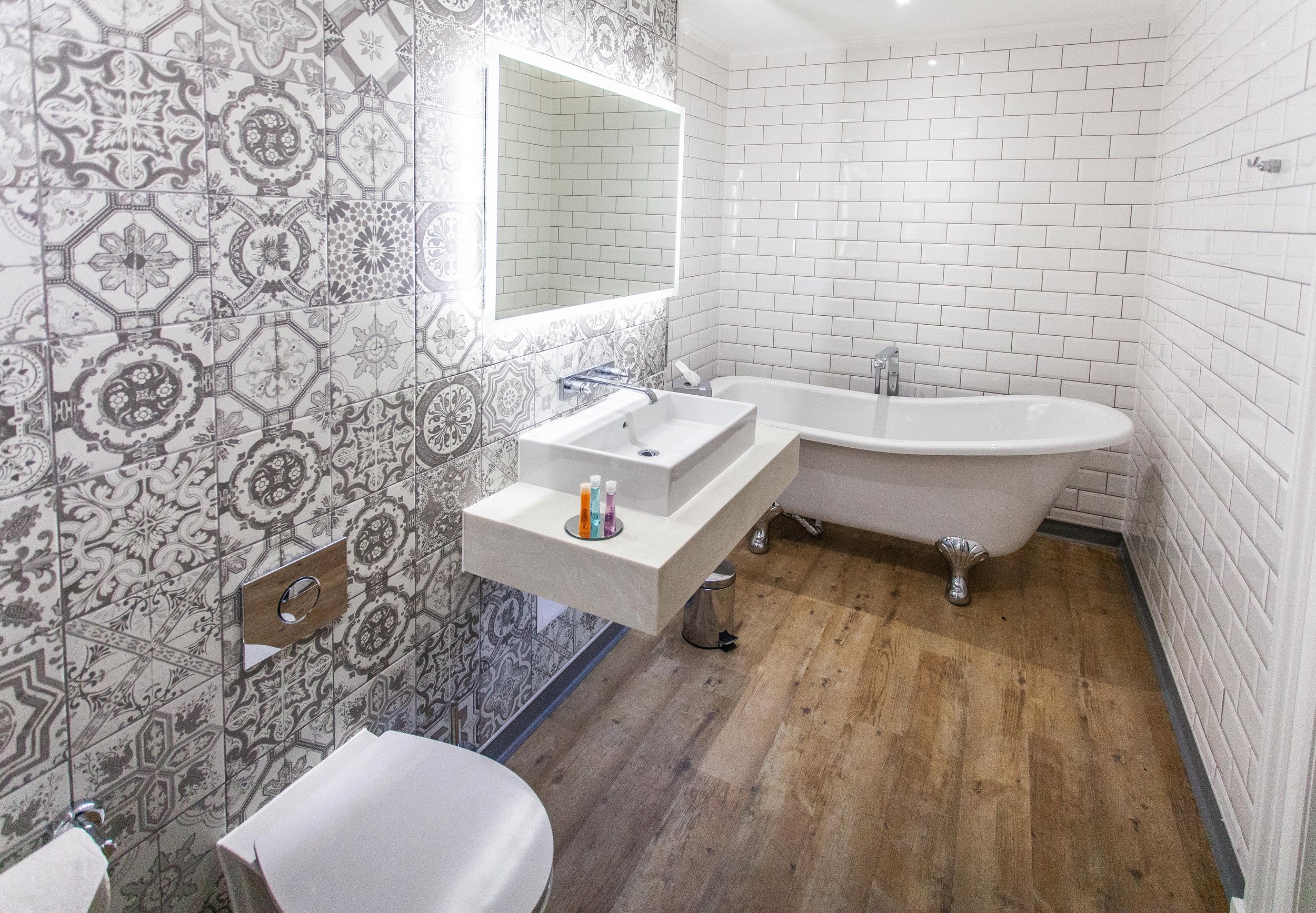 Modern bathroom with decorative patterned wall tiles, a rectangular mirror, a white sink, free-standing bathtub, and wooden flooring.