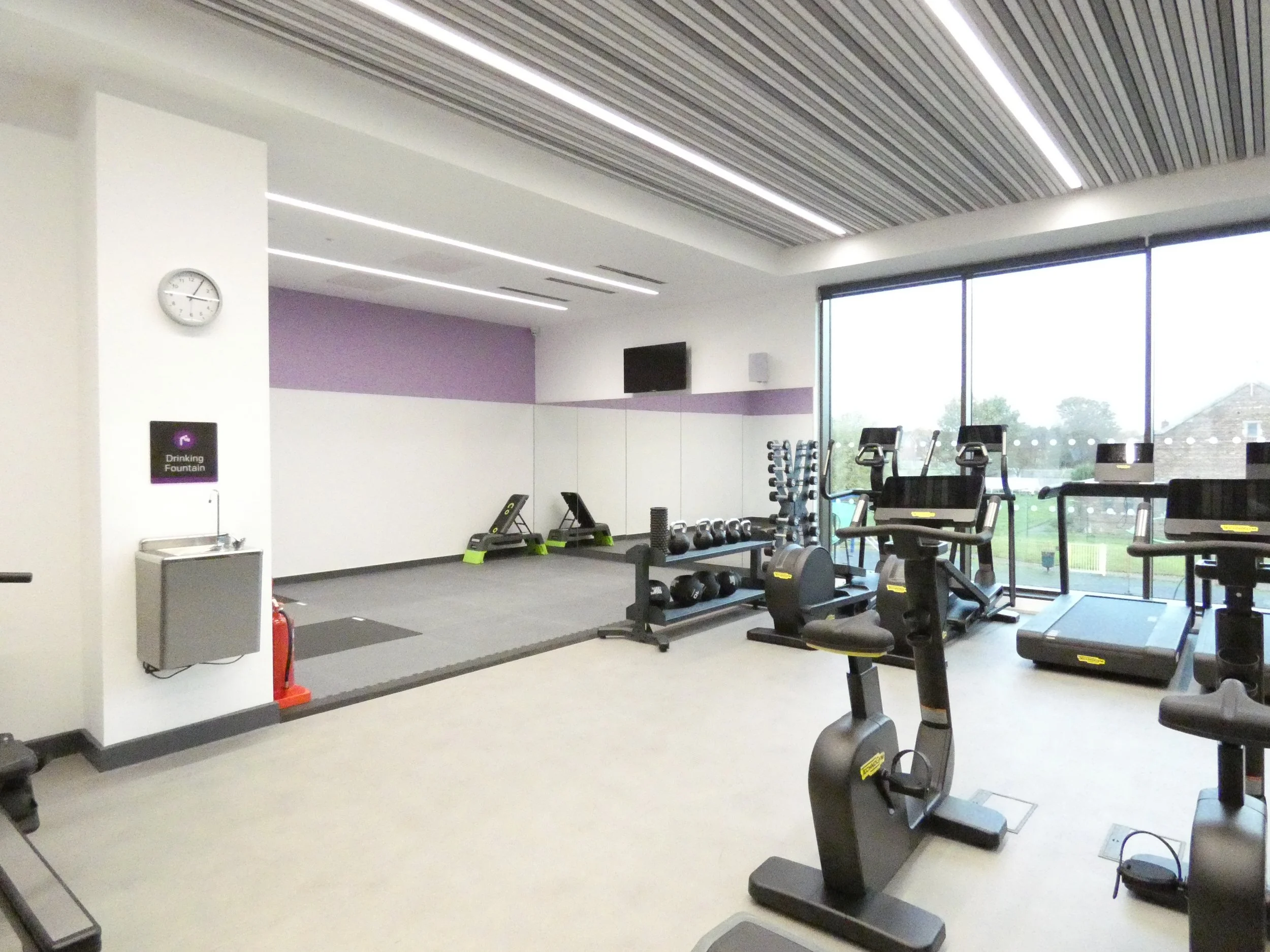 Modern gym with exercise equipment, including stationary bikes, treadmills, and a weight rack, alongside a drinking fountain and clock on the wall.