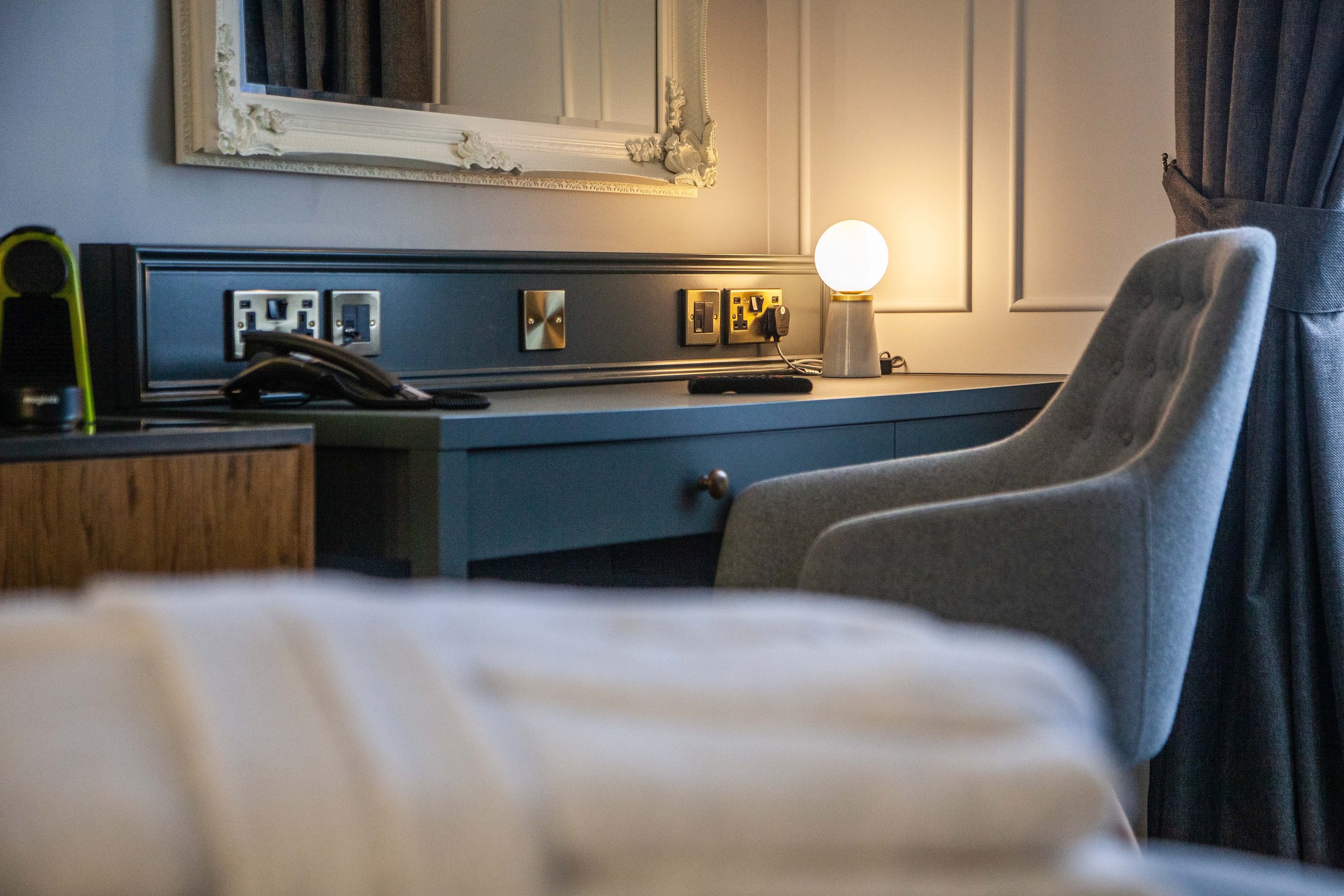 Cozy hotel room desk area with a gray chair, a lamp, a phone, and power outlets. A large mirror hangs above the desk, and a curtain is partially visible.