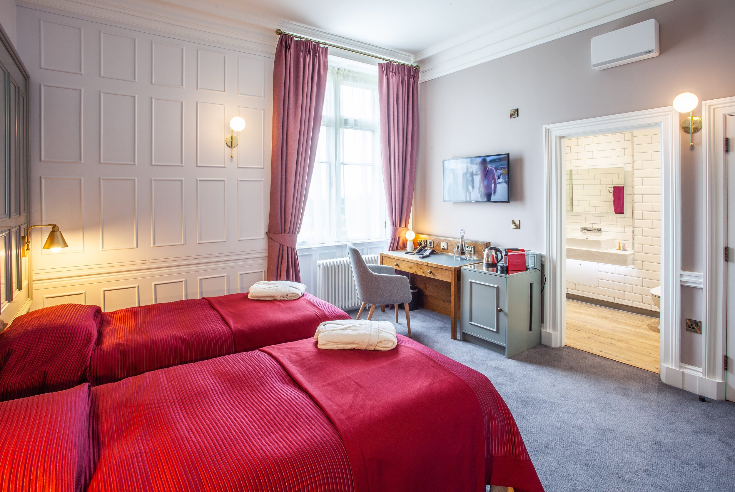 Hotel room with two twin beds, red bedding, wall-mounted TV, desk, chair, kettle, and open door to a white-tiled bathroom.