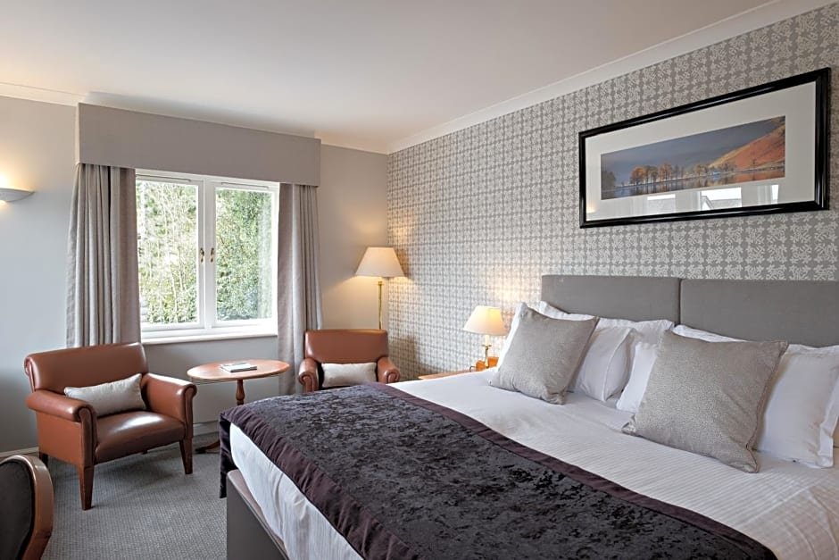 Modern hotel room with a king-sized bed, upholstered headboard, decorative pillows, patterned wallpaper, two leather armchairs, a round wooden table, floor lamp, and a window with curtains.