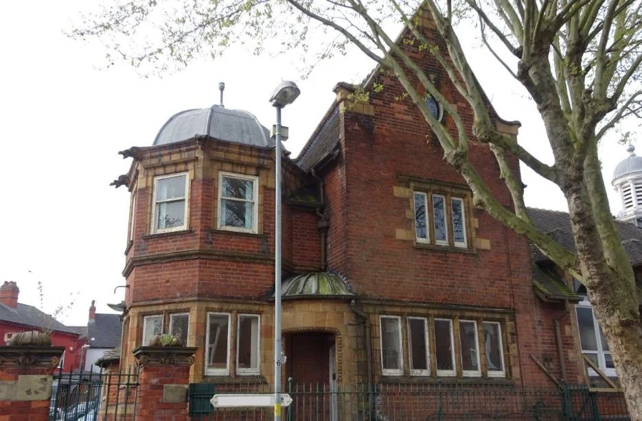 Historic brick building with tower and domed roof surrounded by trees, featuring decorative elements and a metal fence.