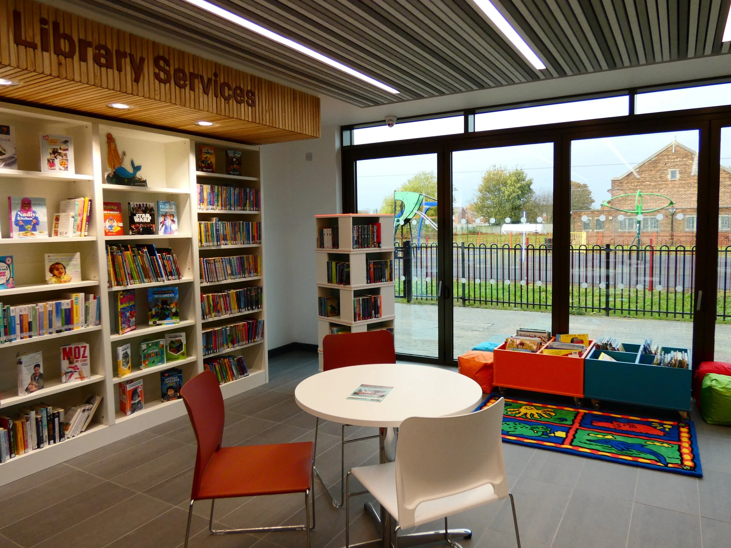 Library interior with children's section featuring bookshelves, a reading table with chairs, a colorful rug, and large windows overlooking an outdoor play area.