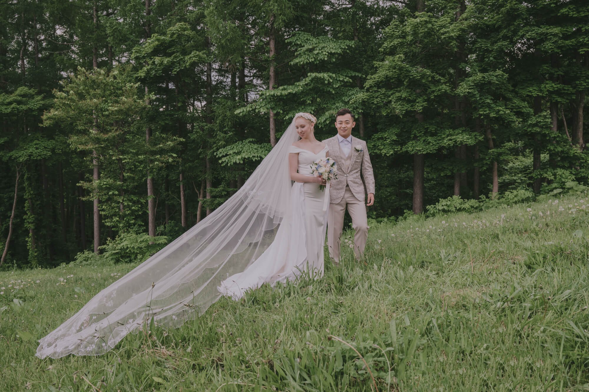 Bride is wedding veil with groom on a grassy hill