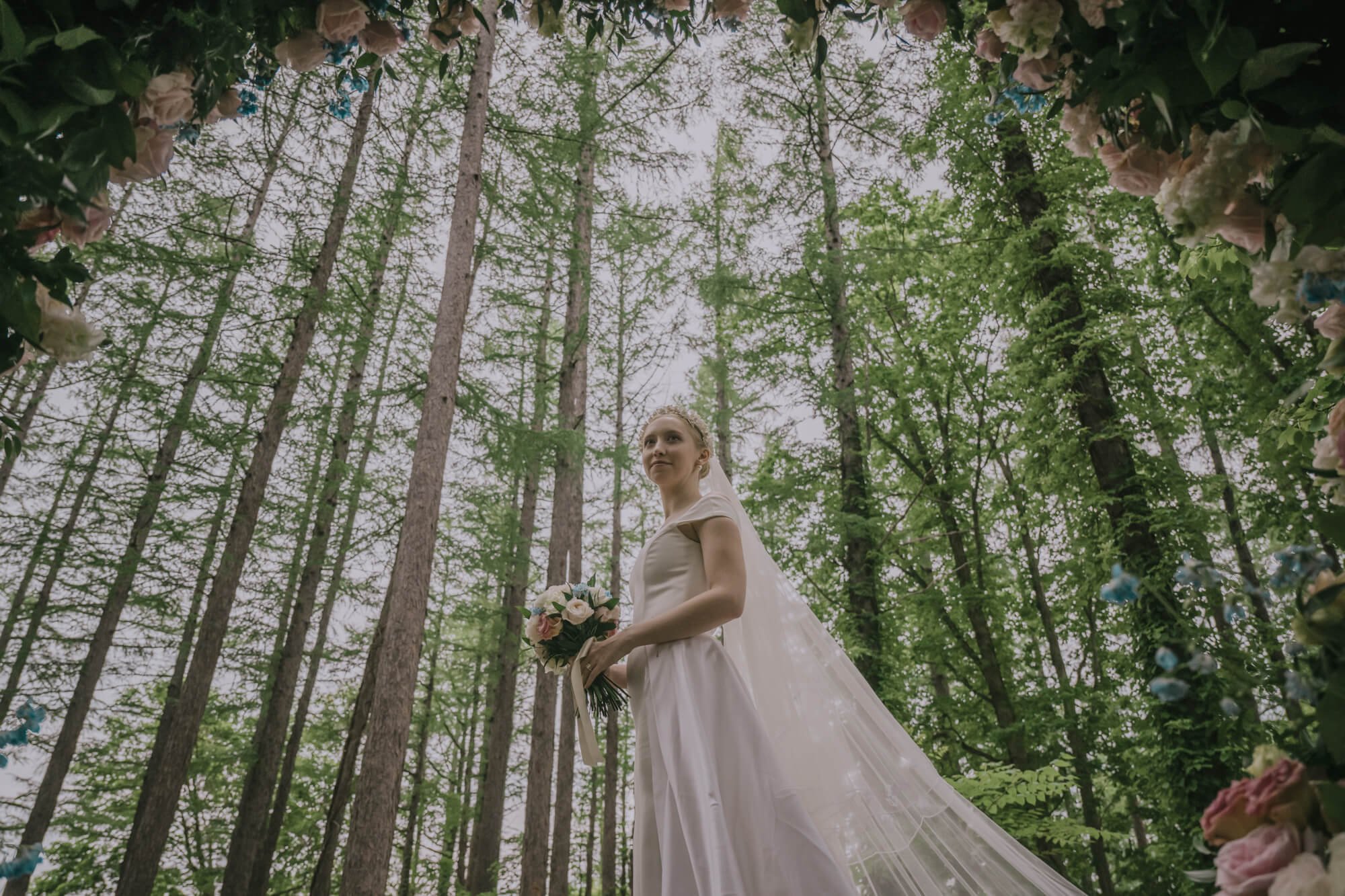 Bride in embroidered veil in front of tall trees