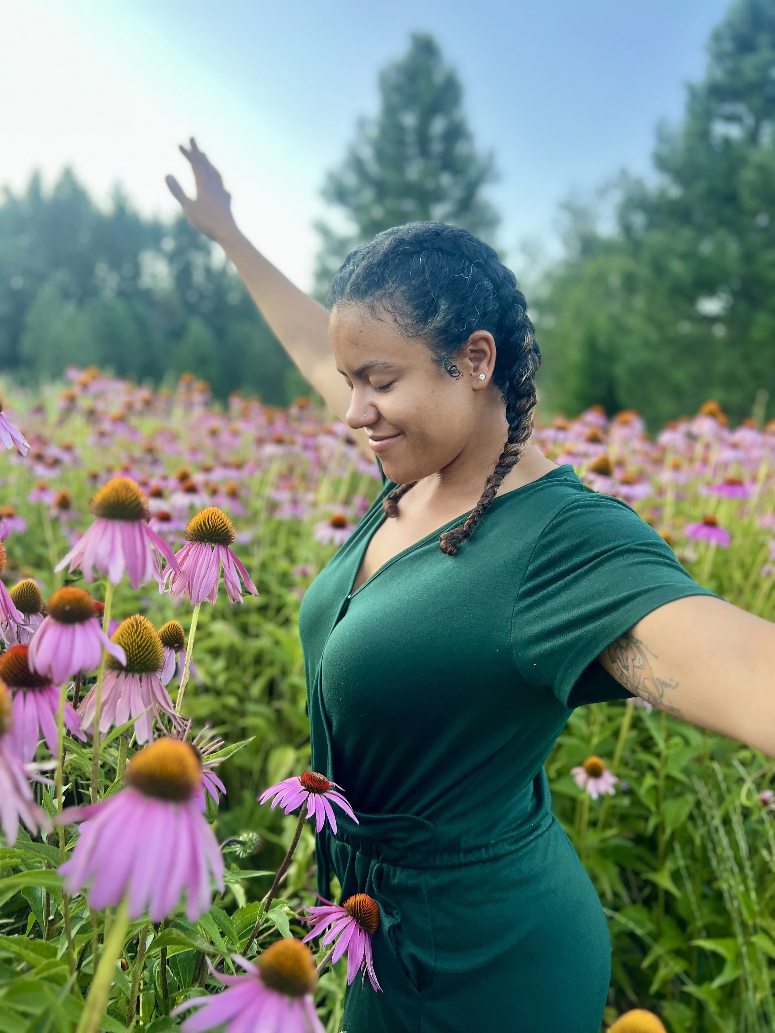 A young woman in a green dress standing among pink coneflowers in a field with her arms outstretched and eyes closed, smiling peacefully.