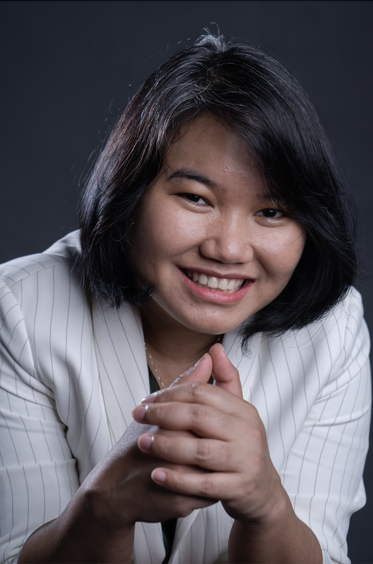A smiling woman with shoulder-length black hair, wearing a white blazer, leaning forward with hands clasped, against a dark background.