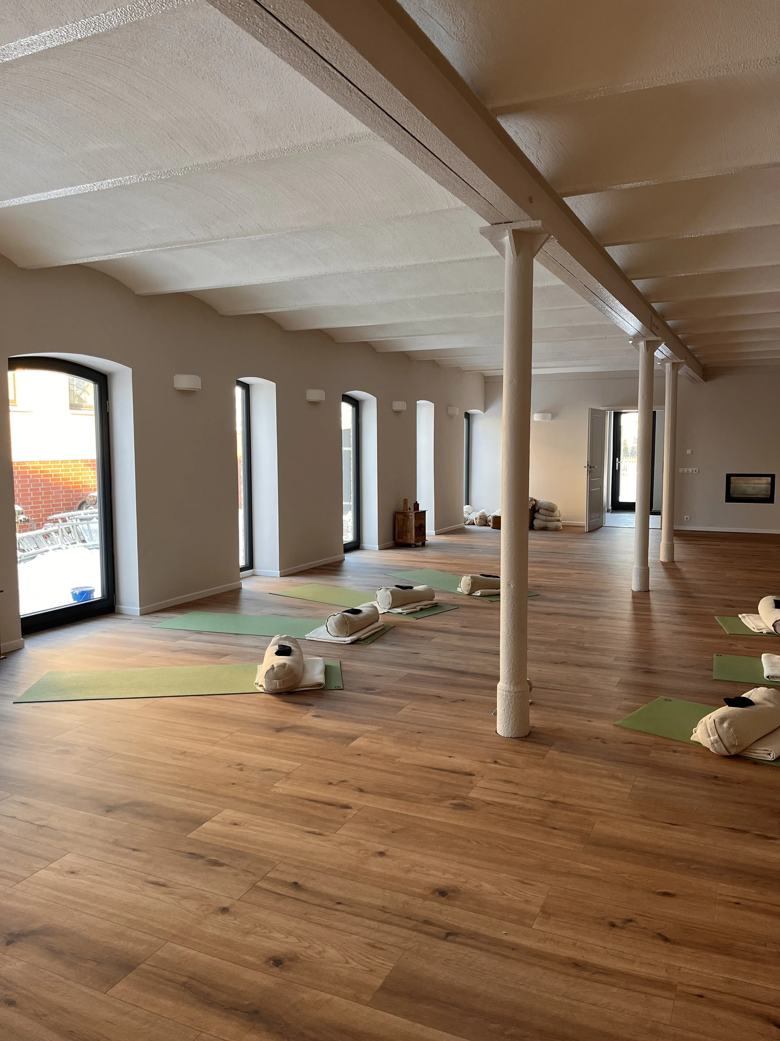 Graue Buddha-Statue mit geschlossenen Augen, umgeben von bunten Blumen in einer goldenen Vase, auf einer Holzscheibe. Gräser im Vordergrund, im Hintergrund ein weißer Zeltstoff.