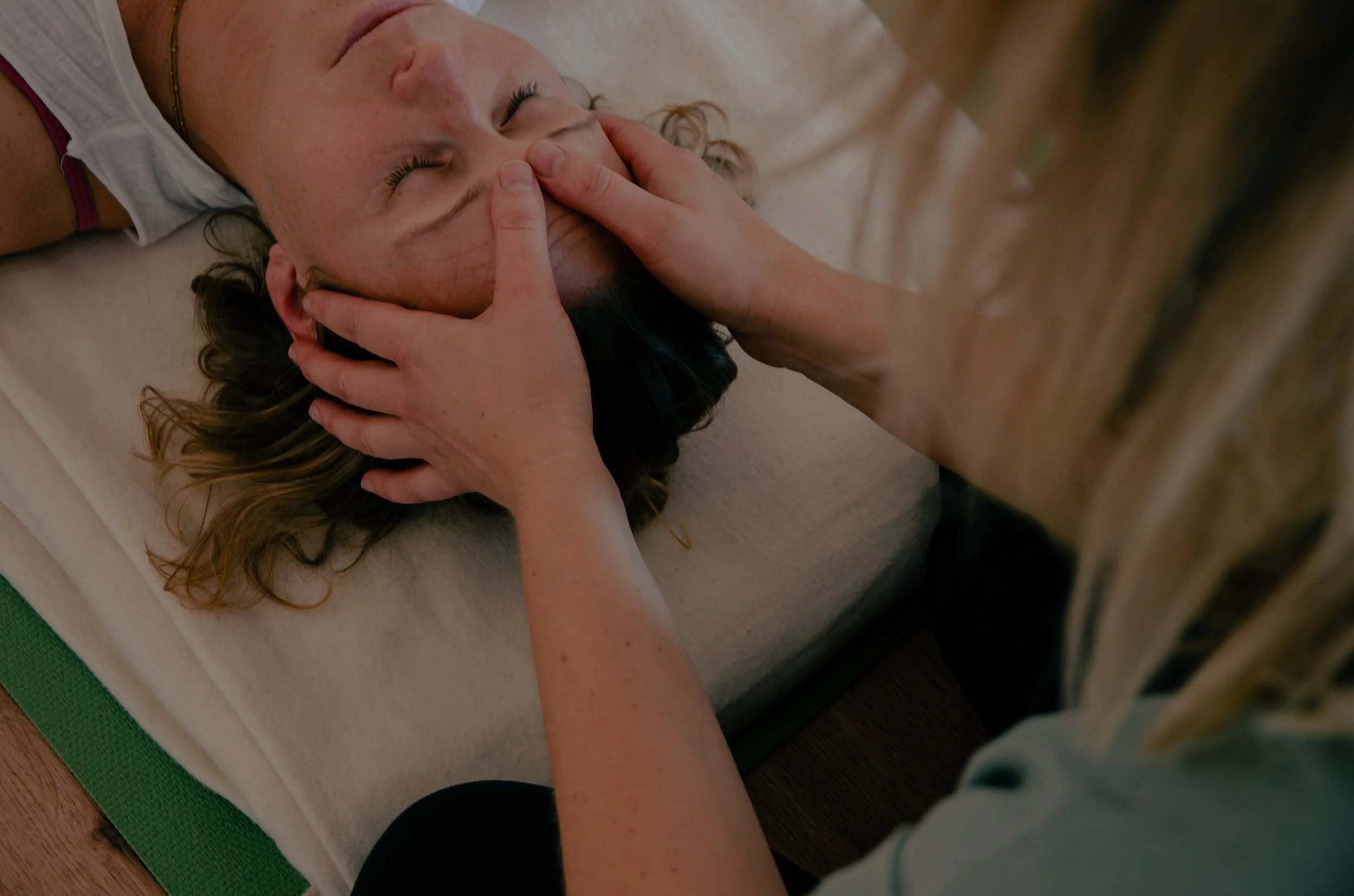 Eine Frau liegt in der Bauchlage auf einer Yogamatte, während eine andere Frau sie im Yoga unterstützt, indem sie ihre Schulter hält und sie in einer Rückenlage hält.
