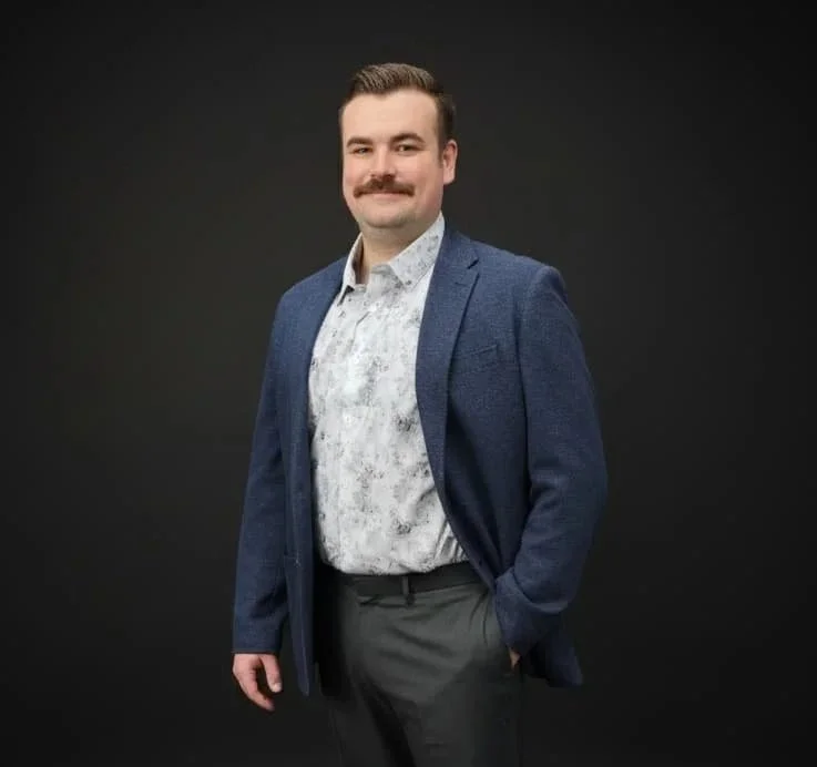 A man with a mustache in a business casual outfit, standing against a dark background, wearing a blue blazer and patterned shirt.