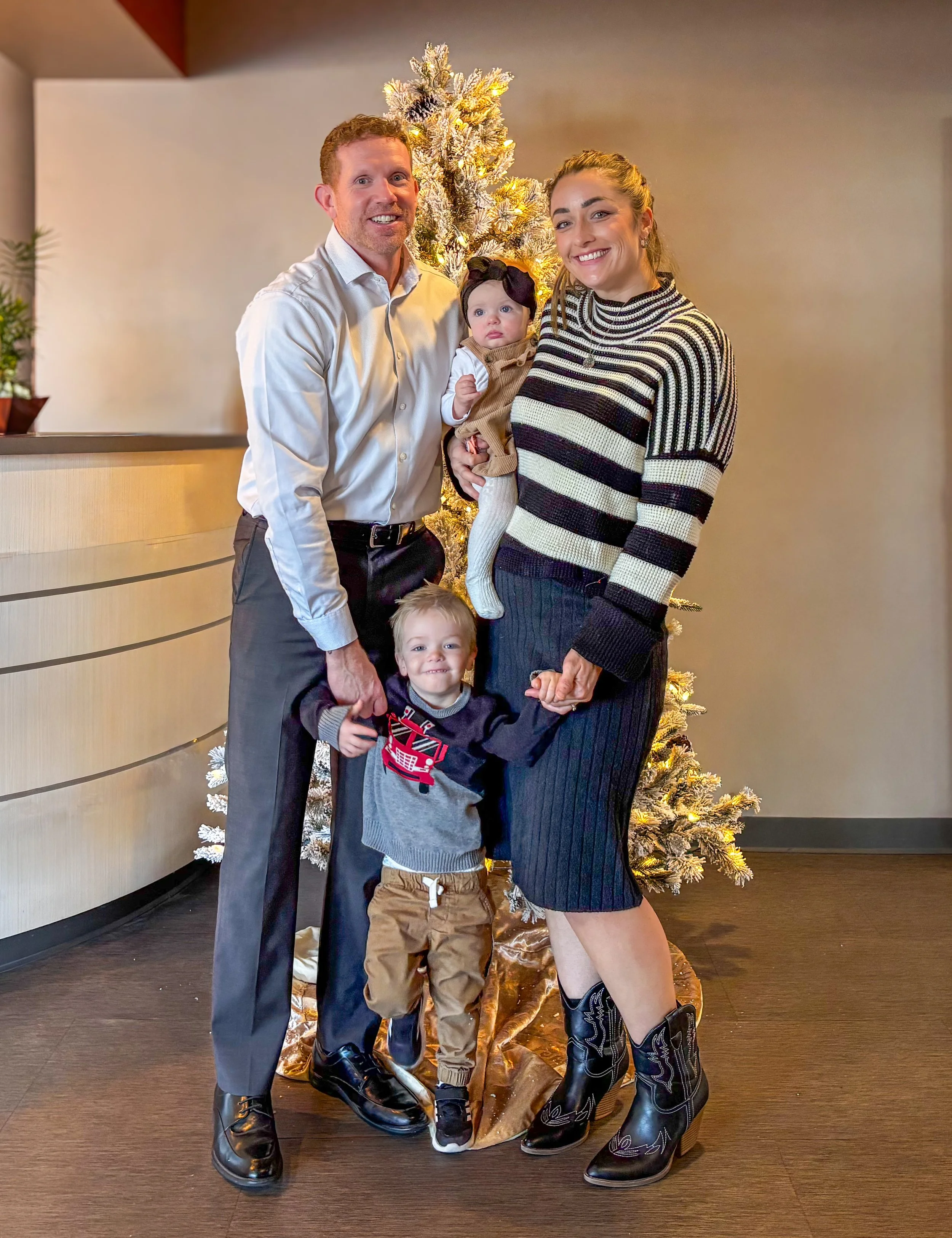 A family of four posing for a photo in front of a decorated Christmas tree indoors. The father has red hair and is wearing a light-colored shirt and dark pants. The mother has long hair tied back, wearing a black and white striped sweater and black s