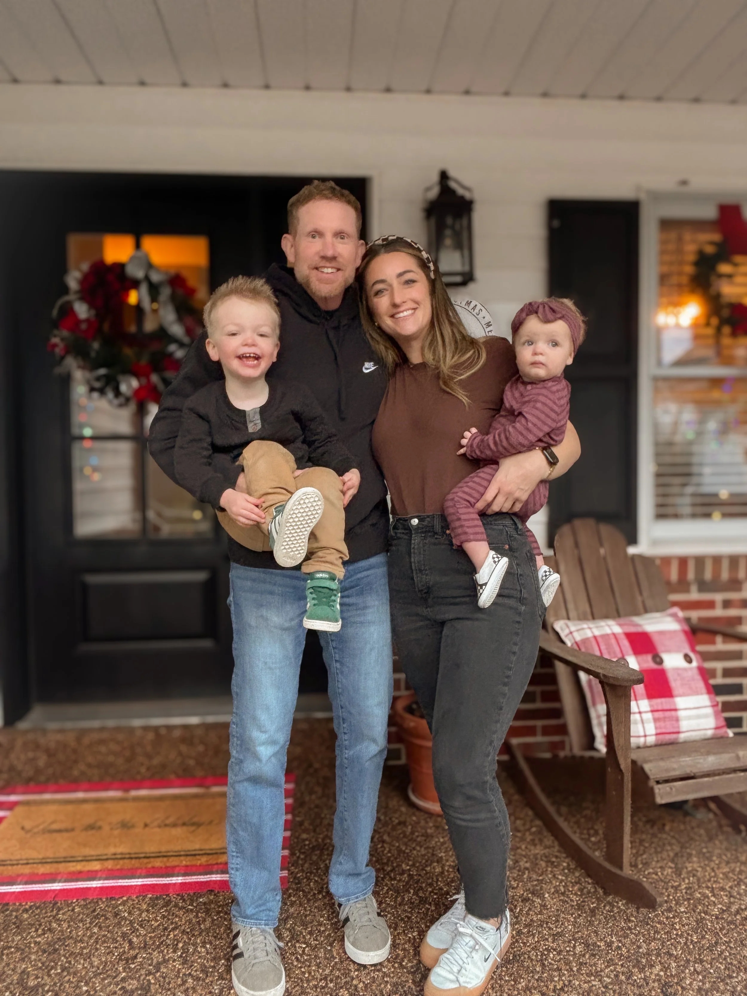 A family of four standing outside on a porch, smiling at the camera, in front of a window decorated with Christmas wreaths and lights.