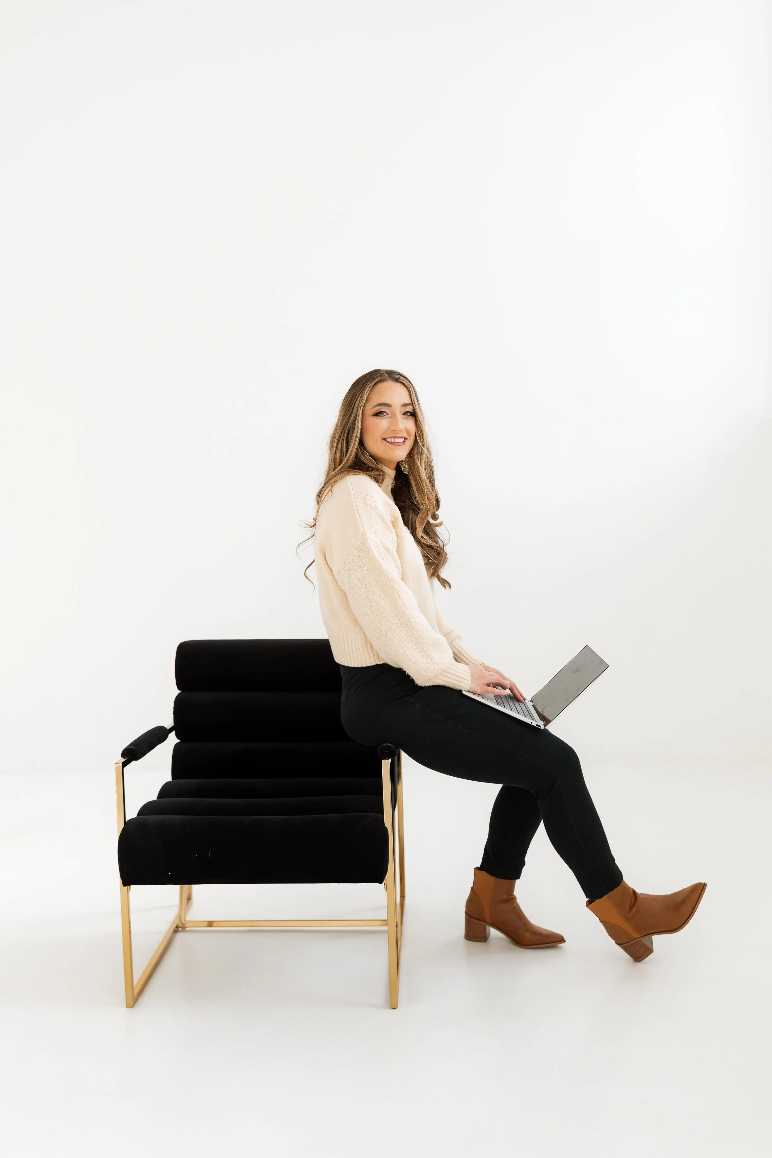 A woman with long wavy hair, dressed in a cream sweater and black pants, sitting on a black chair and using a laptop, in a minimal white studio setting with a simple chair behind her.