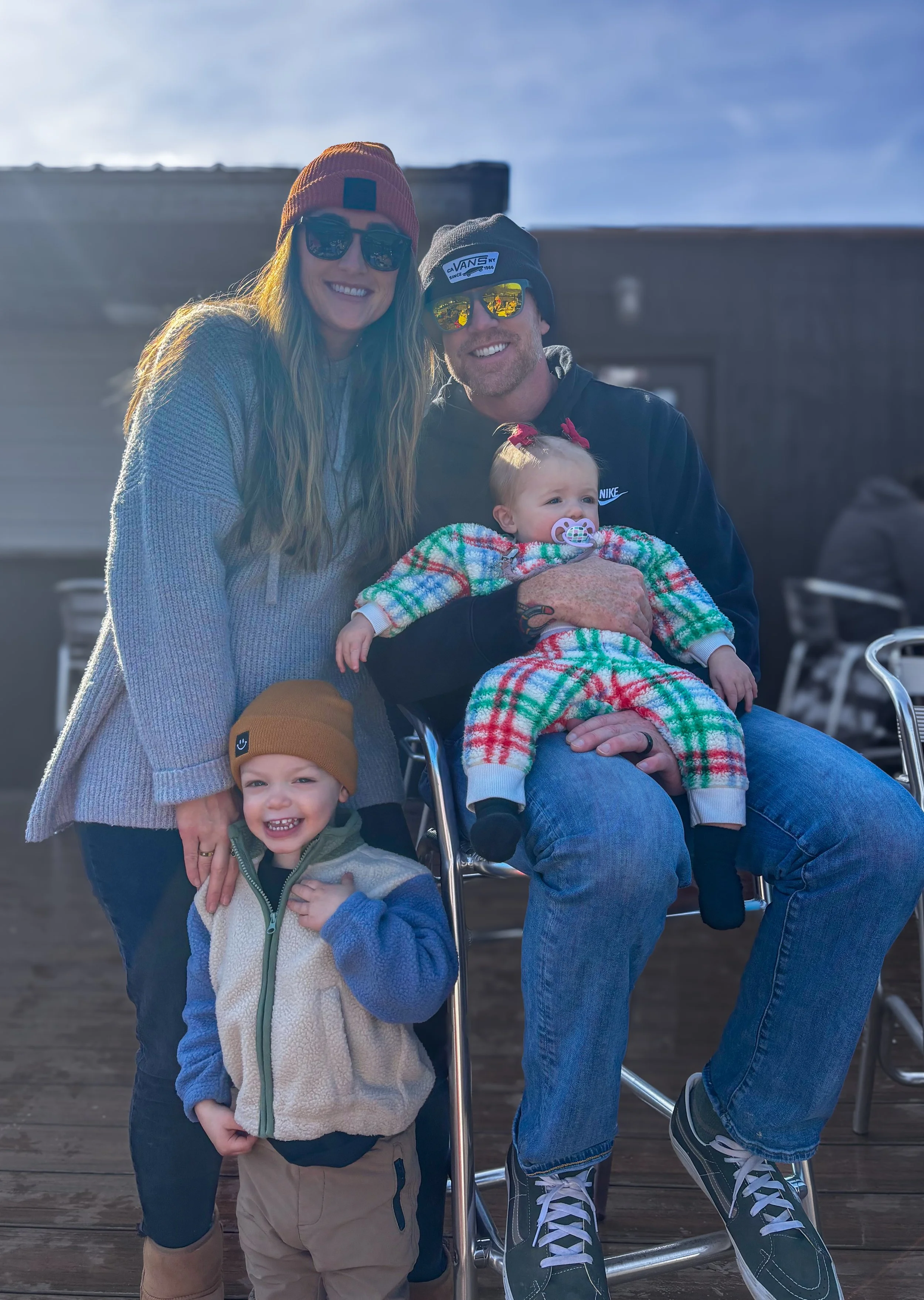 Happy family of four outdoors on a sunny day, with a woman, man, young boy, and baby girl, smiling and posing for the photo.