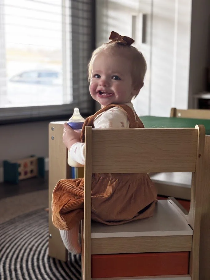 A smiling baby girl with a bow headband sitting on a small wooden chair at a table, holding a baby bottle, near a window in a cozy room.