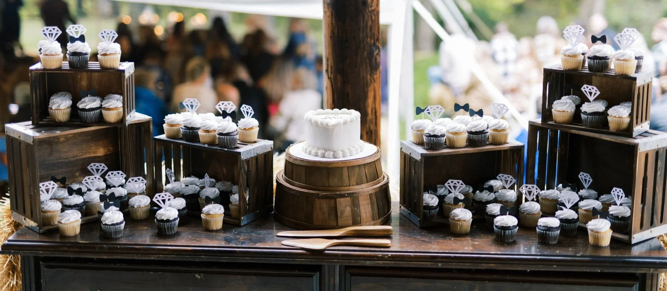 Cupcakes with diamond and bowtie decorations arranged on wooden crates and barrels at an outdoor celebration.