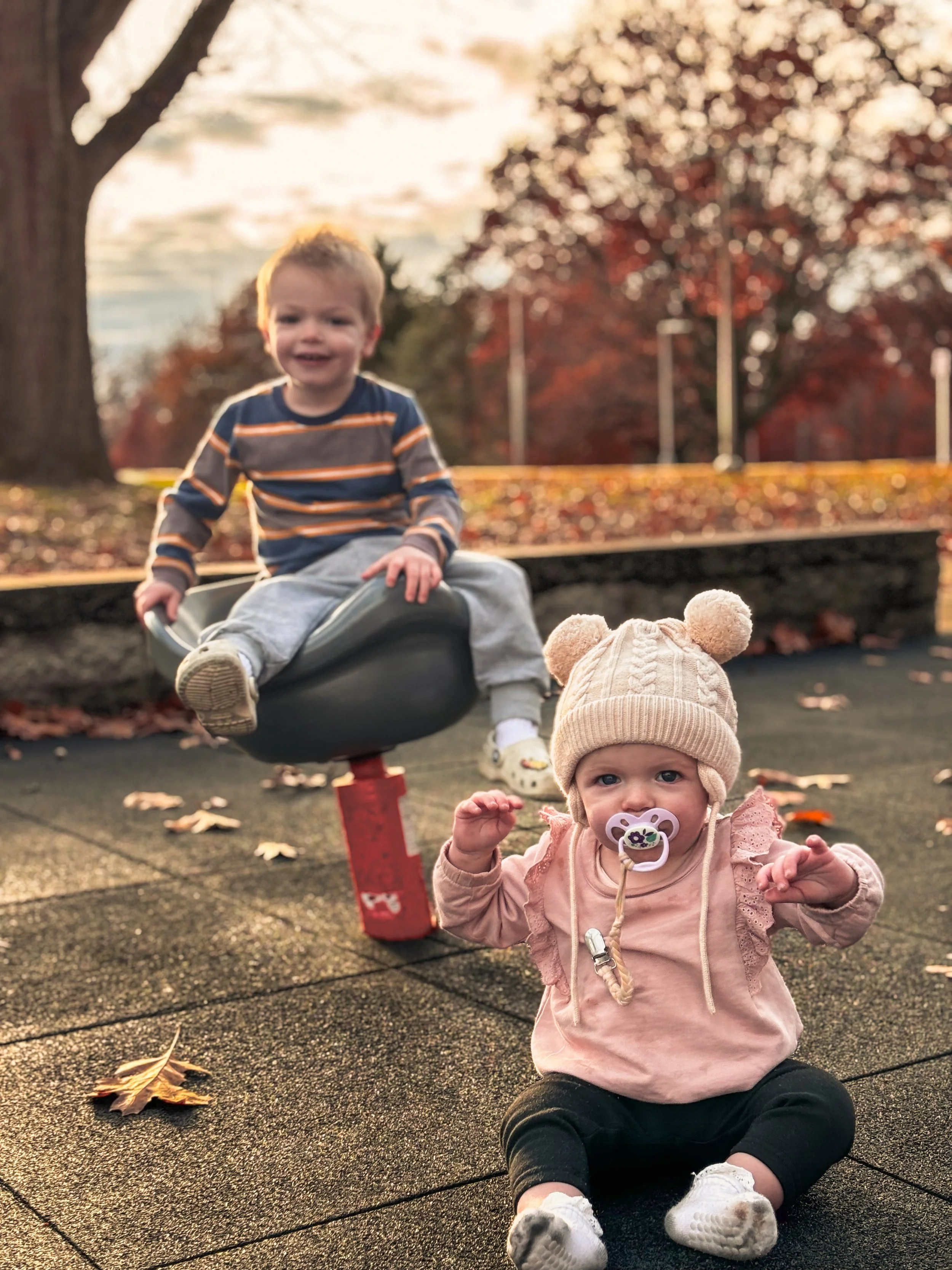 Two babies playing outdoors on a sunny autumn day. One baby is sitting on the ground wearing a hat and pacifier, and the other is smiling and playing on a seesaw in the background.