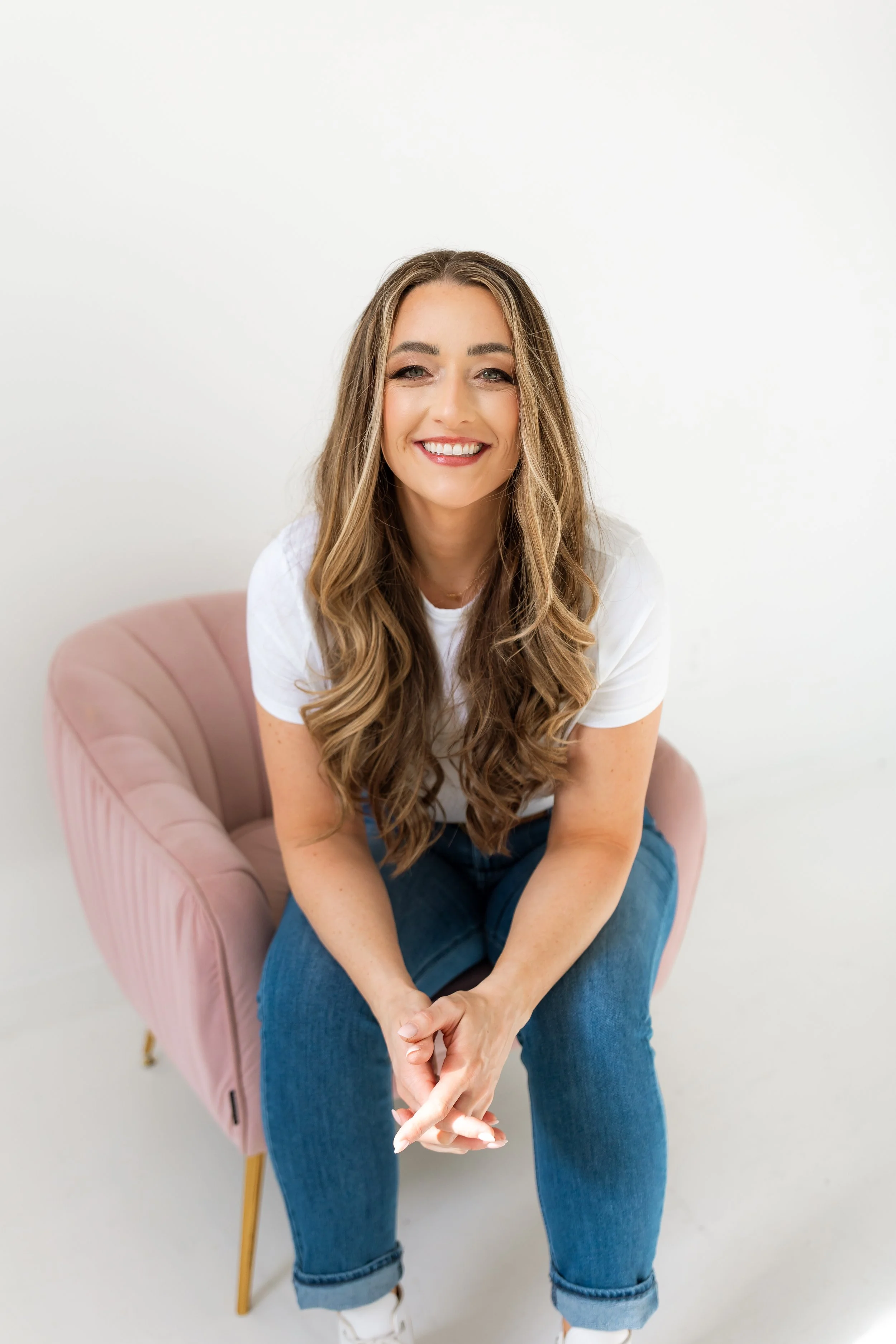 A young woman with long wavy hair, wearing a white t-shirt and blue jeans, sitting on a pink armchair, smiling at the camera against a white background.