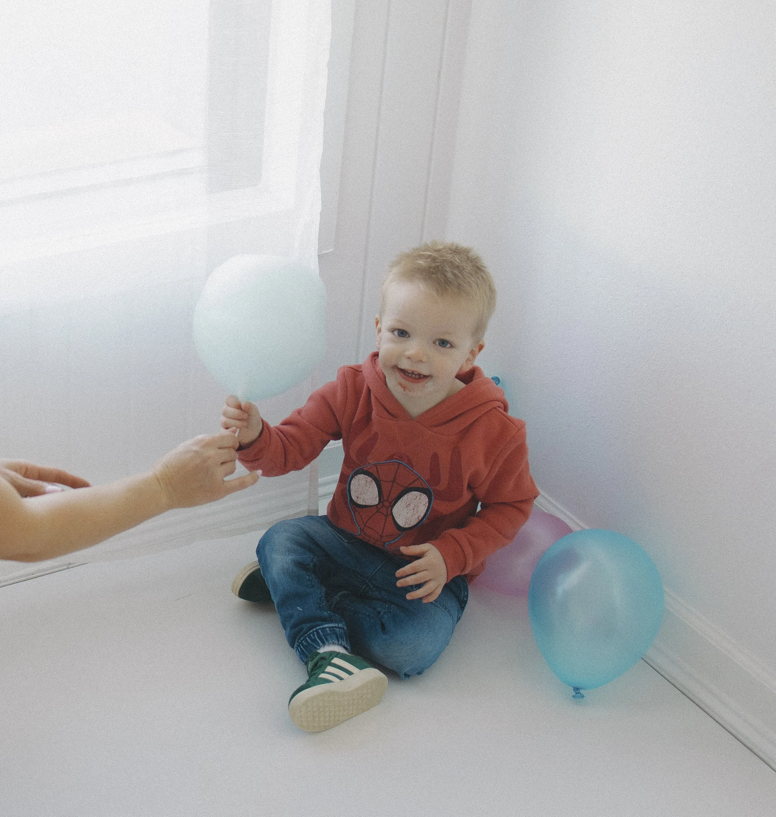 A young boy with red hair, wearing a red hoodie with a Spider-Man logo, sitting on a white floor surrounded by balloons, smiling and holding a balloon.
