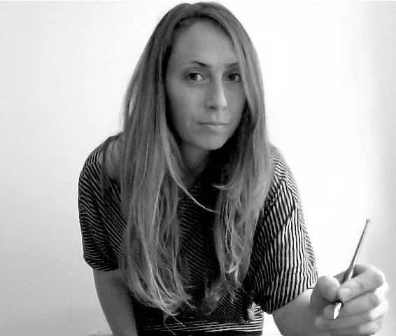 Black and white photo of a woman with long hair, wearing a striped shirt, holding a pencil, and looking at the camera.