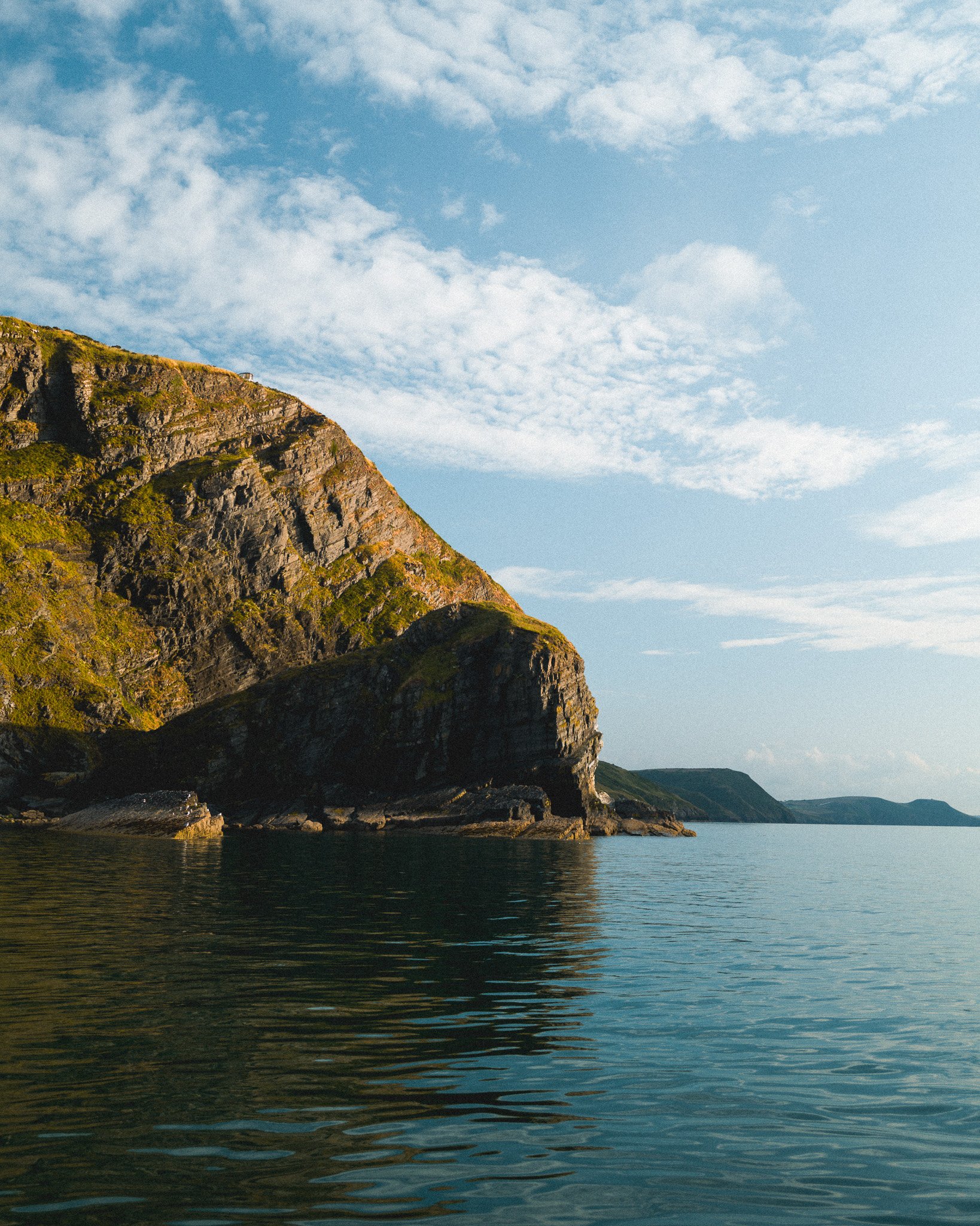 A rocky coastline with steep cliffs covered in green vegetation next to calm water under a partly cloudy sky.
