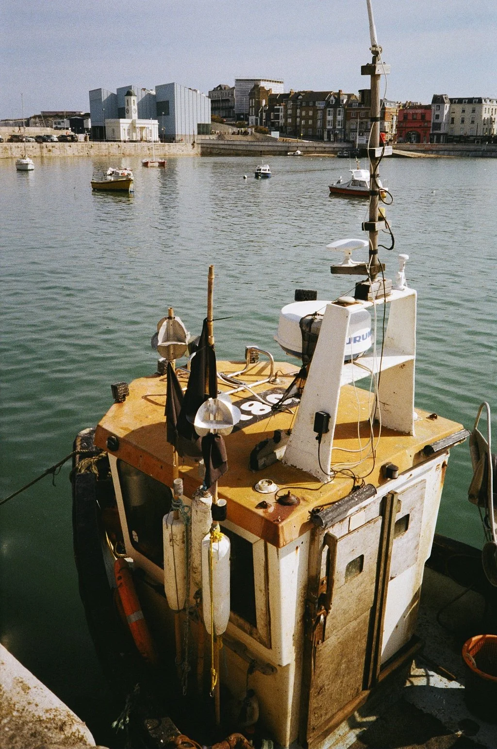 A small, weathered boat docked at a harbor with several other boats floating in the water and a cityscape with modern and historic buildings in the background.