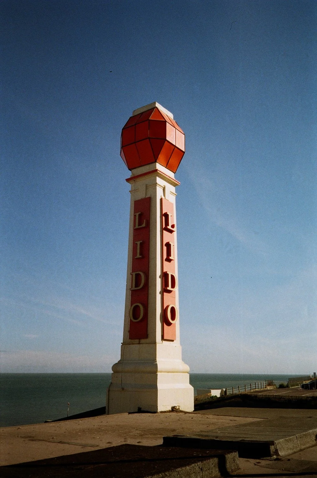 A lighthouse with a red, geometric-patterned top and the word 'LIDO' vertically in red on a white column, overlooking the ocean under a clear blue sky.