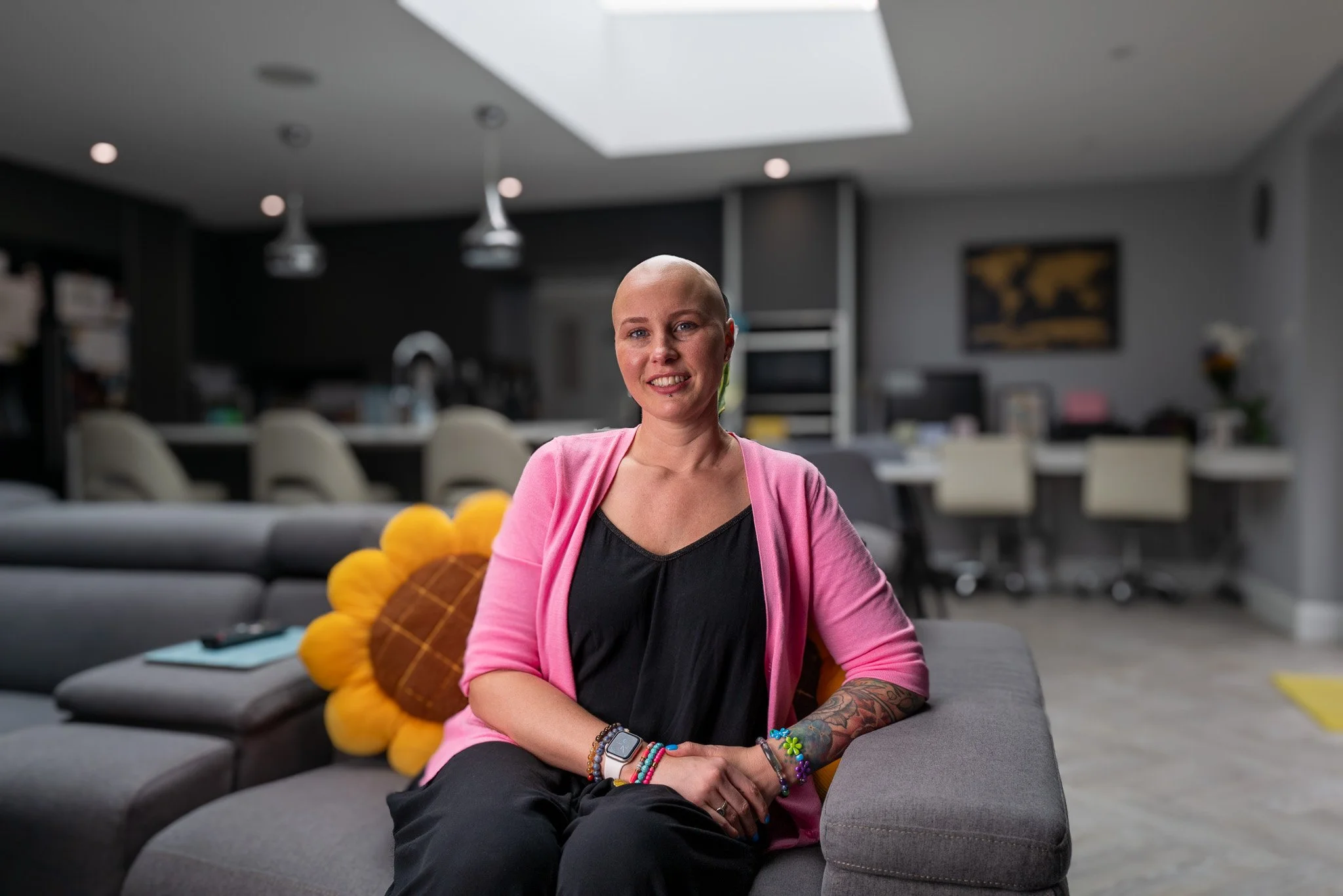 A woman with a shaved head sitting on a gray sofa in a modern, open-plan living room and kitchen area, smiling at the camera, with colorful bracelets and tattoos on her arm, a sunflower-shaped pillow behind her.