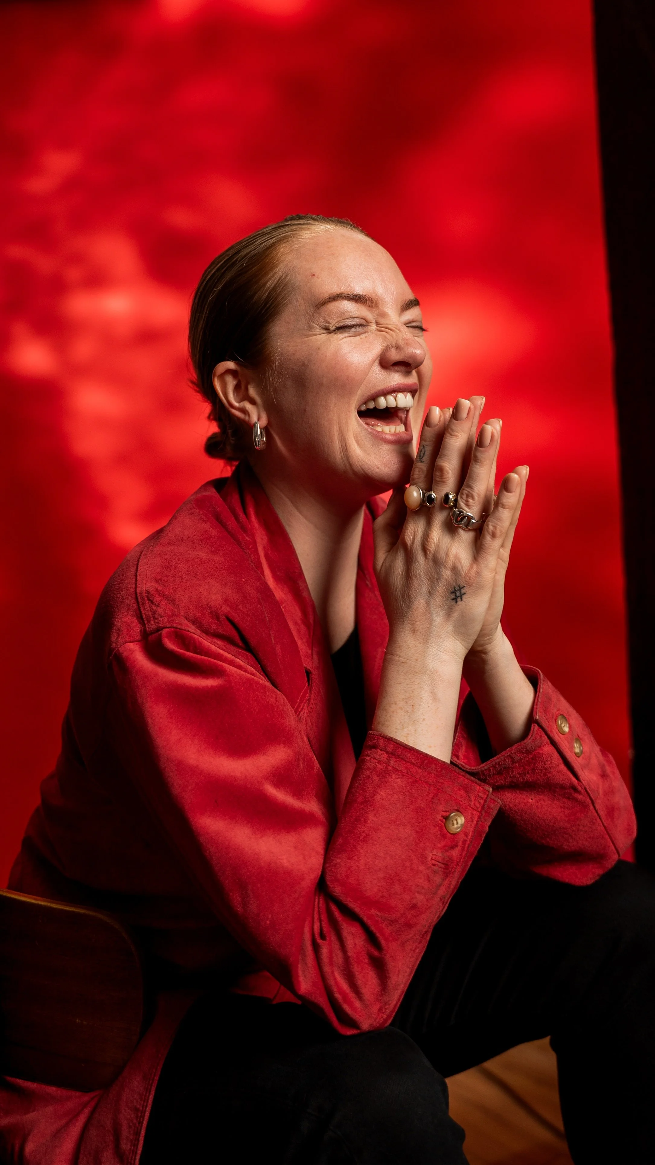 A woman with short brown hair, wearing a red jacket, laughing joyfully with her eyes closed, sitting on a chair against a red background.
