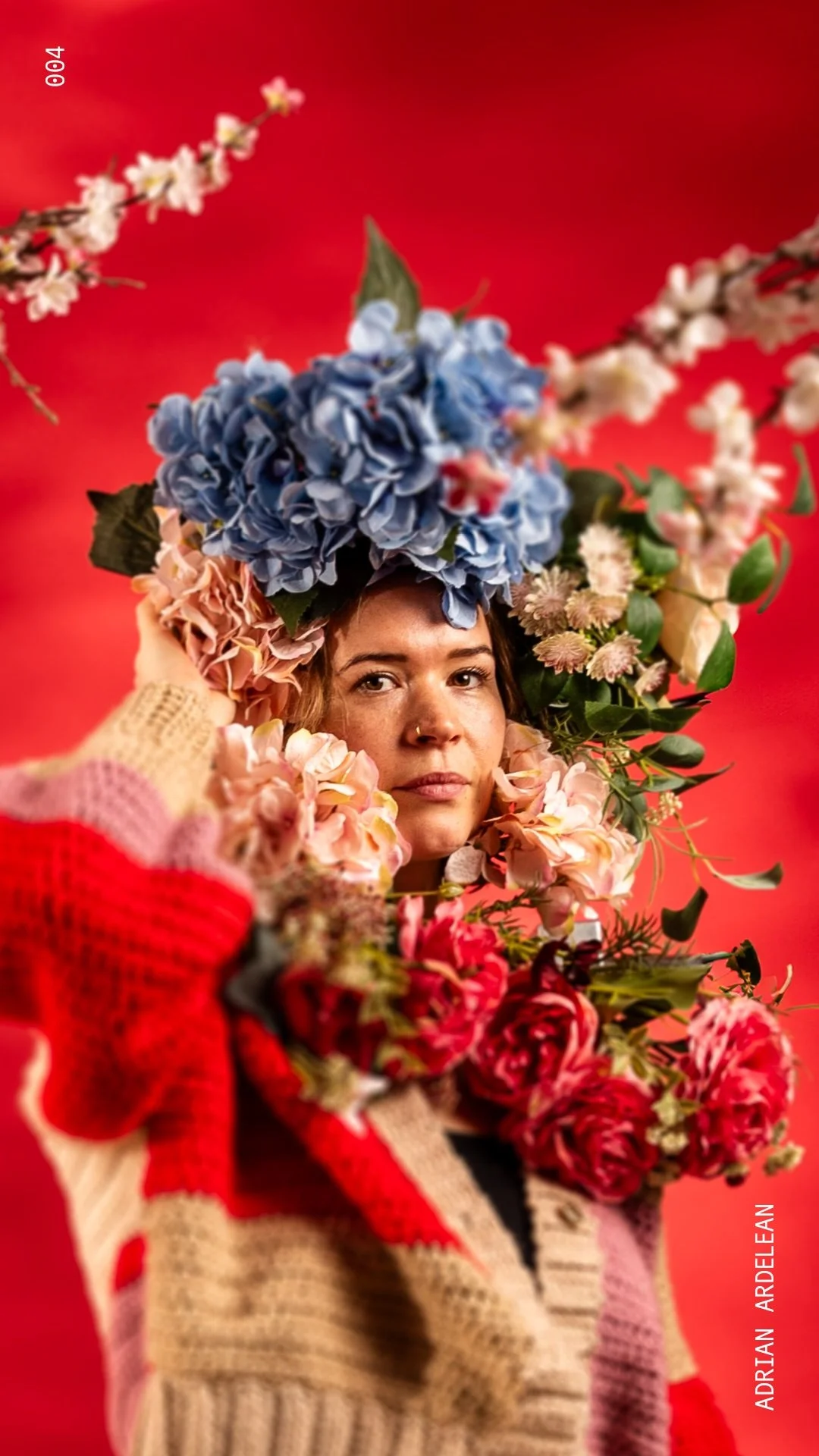 A woman with a serious expression surrounded by colorful flowers, holding flowers near her face against a red background.